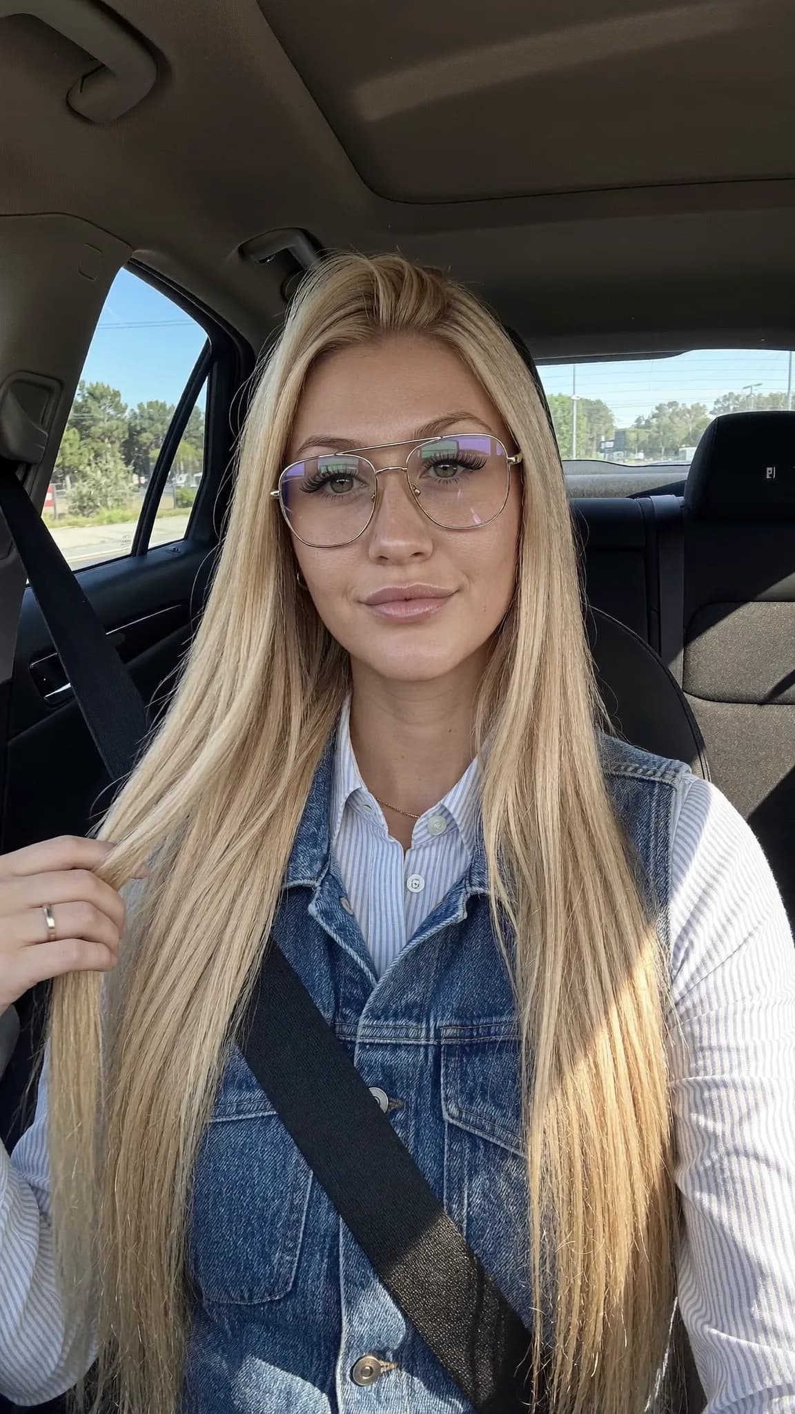 A woman with long blonde hair and aviator glasses is sitting in a car, wearing casual clothing.
