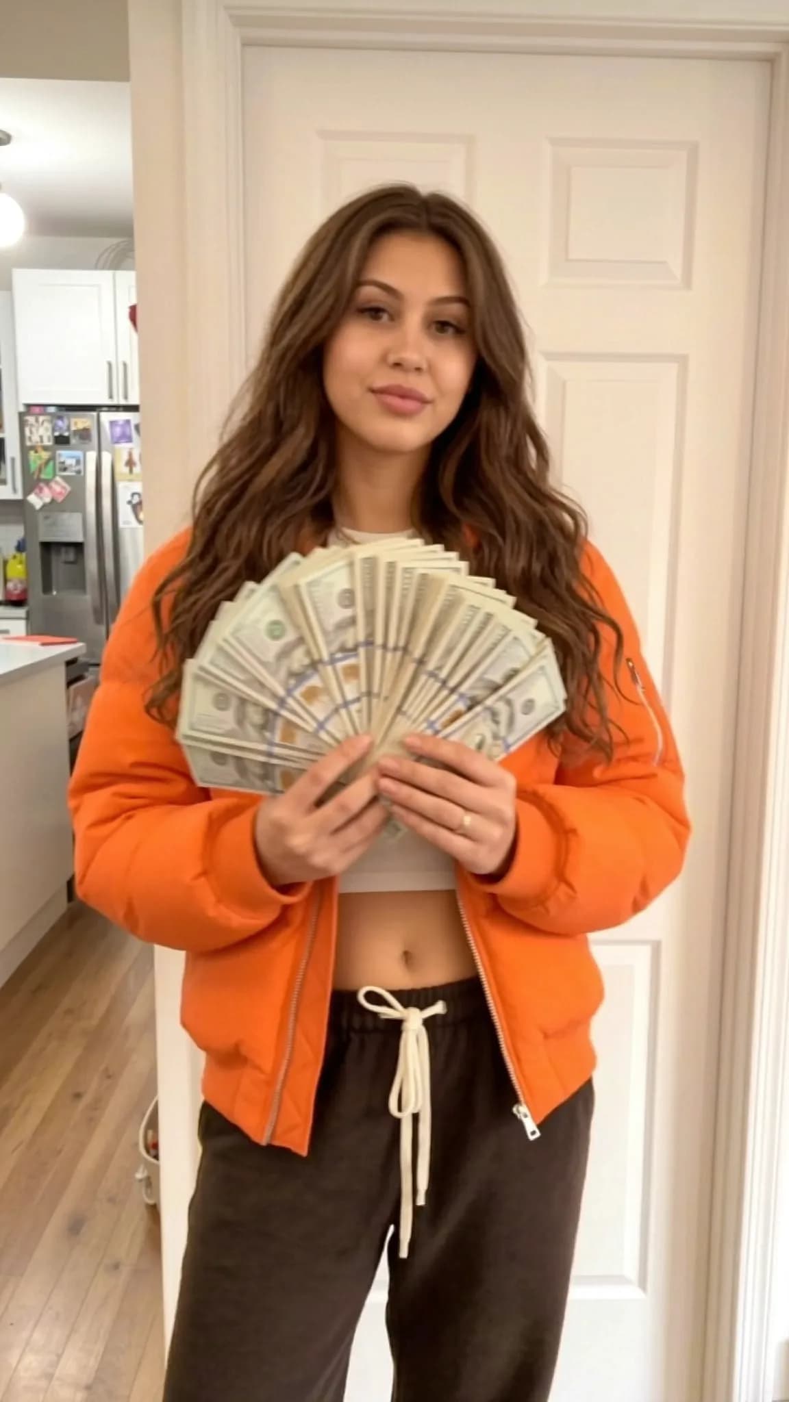 A person in an orange jacket holds a large stack of cash while standing in a kitchen.