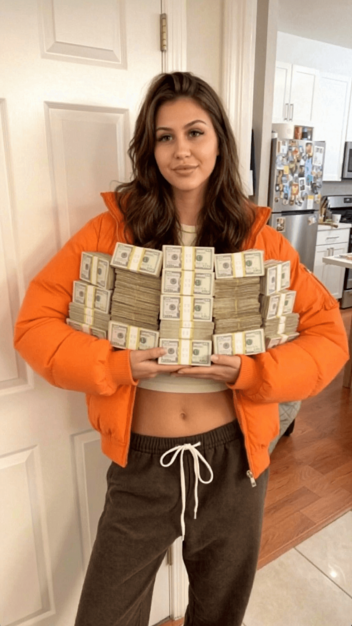 A person in casual attire holds a large stack of cash while standing in a kitchen.