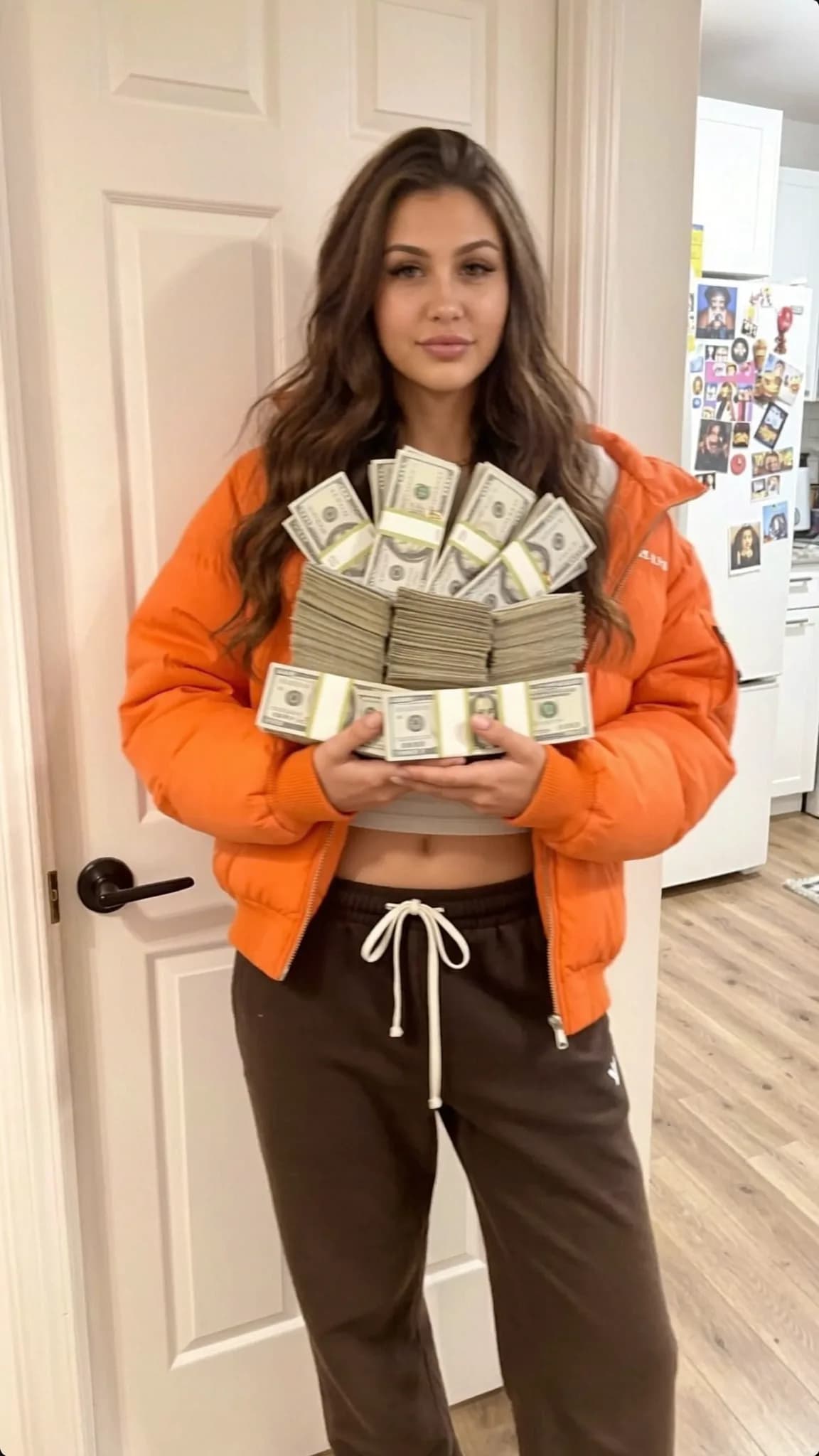 A person in an orange jacket holds a large stack of cash while standing in a kitchen.