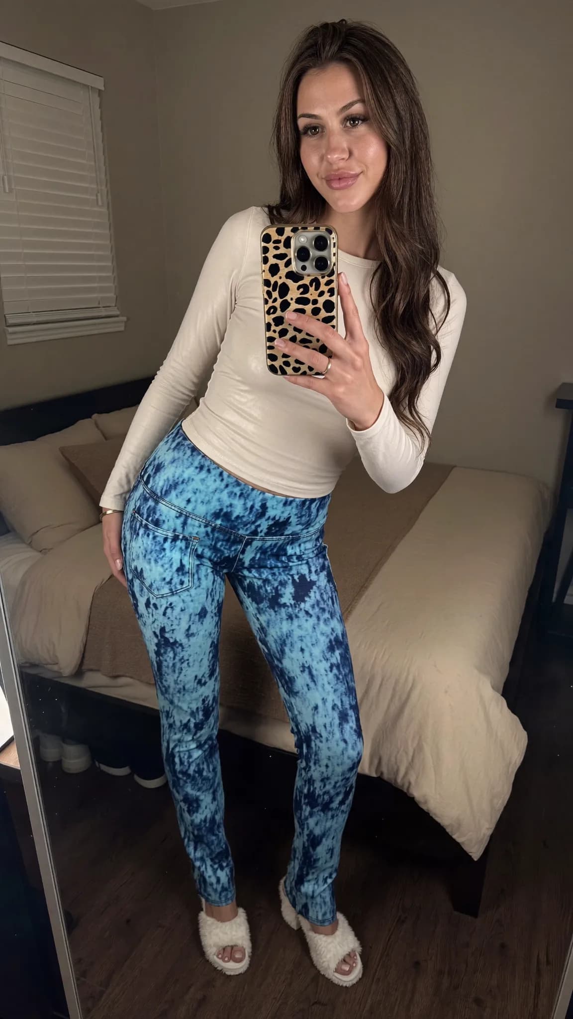 A woman in casual attire poses for a mirror selfie in her bedroom.