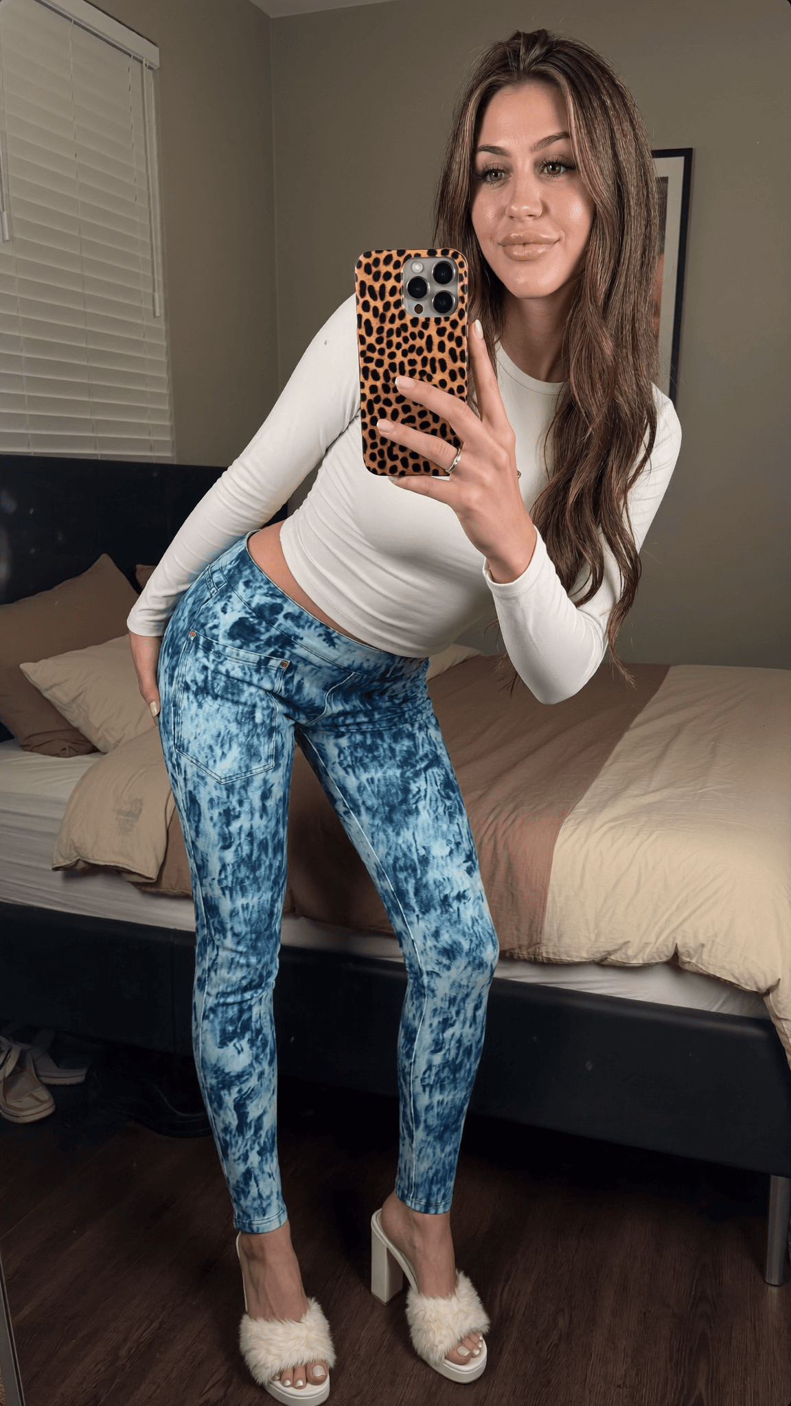 A woman in a bedroom poses with a leopard phone case, wearing blue tie-dye pants and white heels.