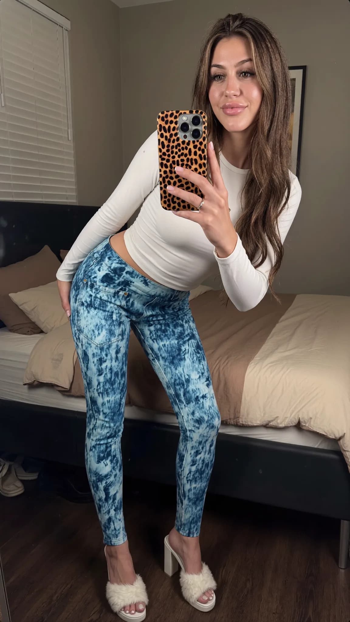 A woman in a bedroom poses with a leopard phone case, wearing blue tie-dye pants and white heels.