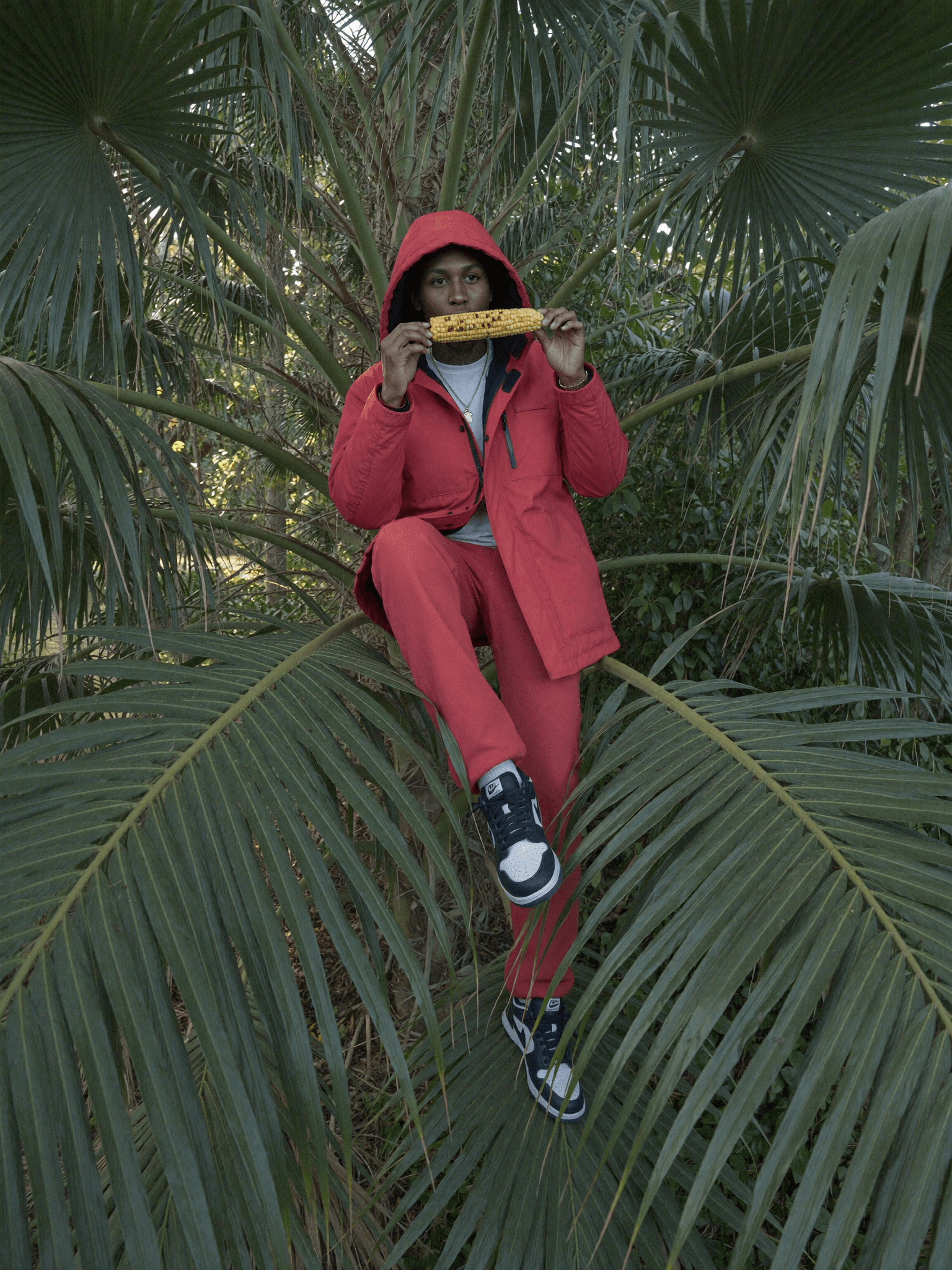 A person in a red outfit sits on palm leaves, holding corn and smiling.