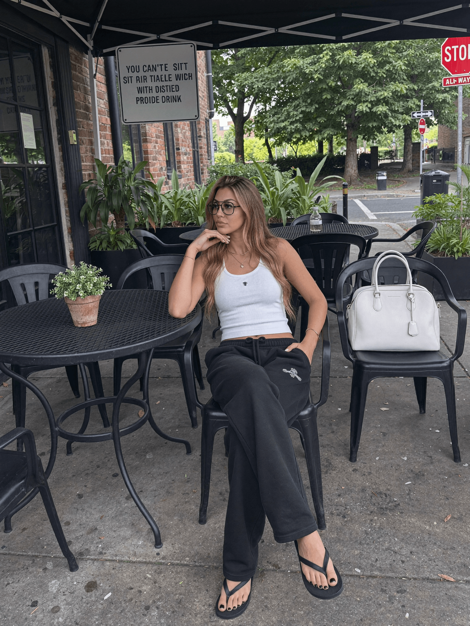 A woman in casual athleisure sits at an outdoor café table with a white handbag on the chair next to her.