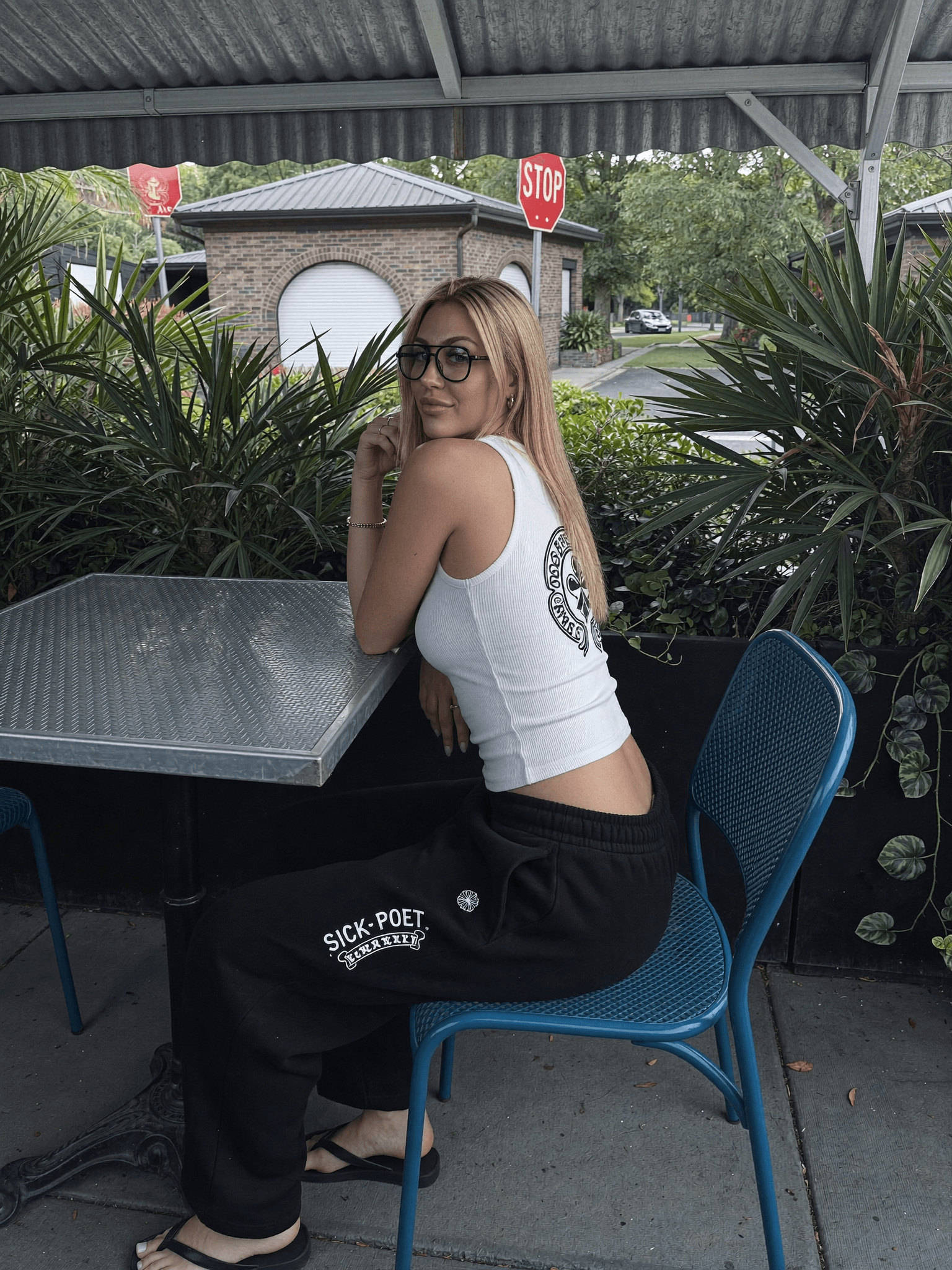 A woman in a white tank top and black sweatpants sits at an outdoor table, with greenery and stop signs visible in the background.