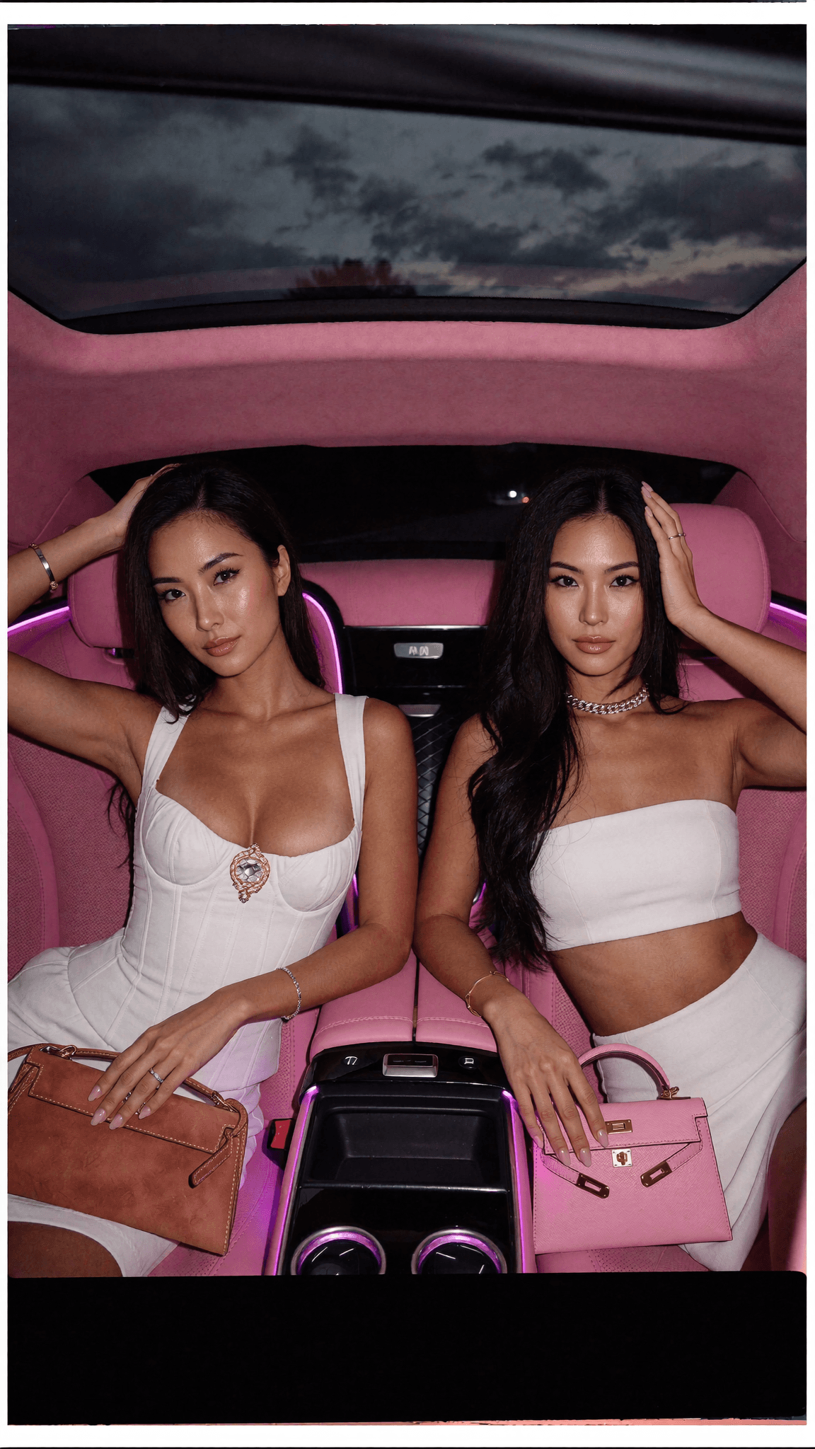 Two women in white dresses sit inside a pink car, posing for the camera.
