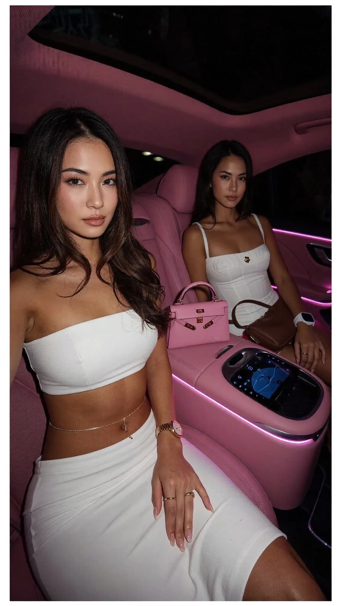 Two women in white dresses sit inside a pink car, exuding confidence and style.
