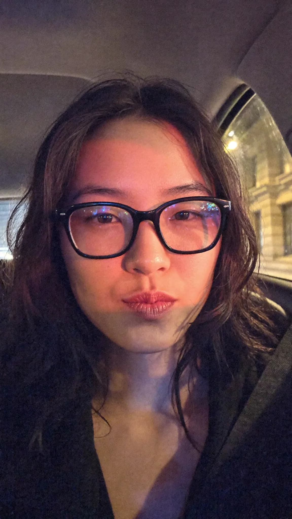A person with long dark hair and glasses is sitting in a car, wearing casual clothing.