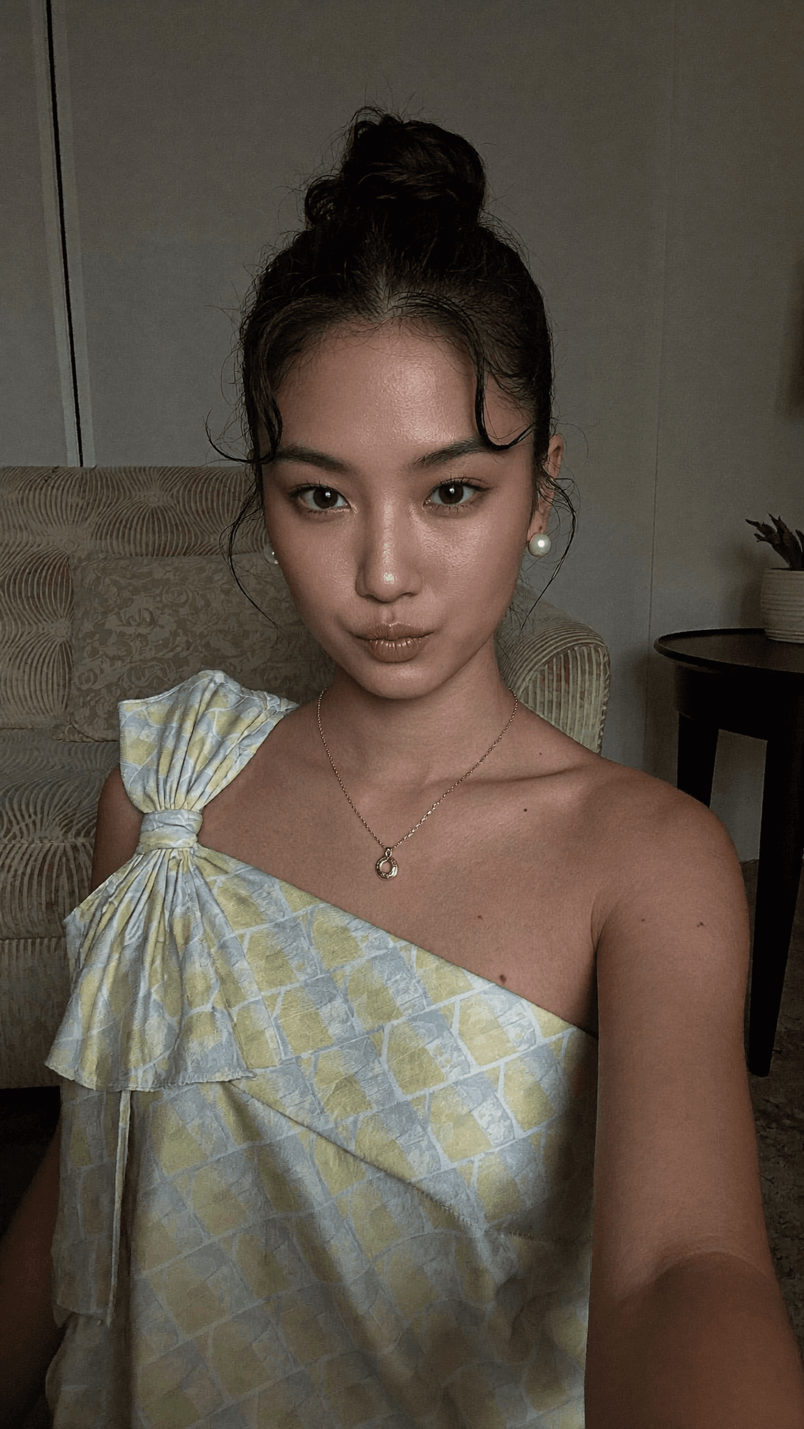 A woman with a pixie cut and beauty mark poses in a bedroom, wearing a dress.