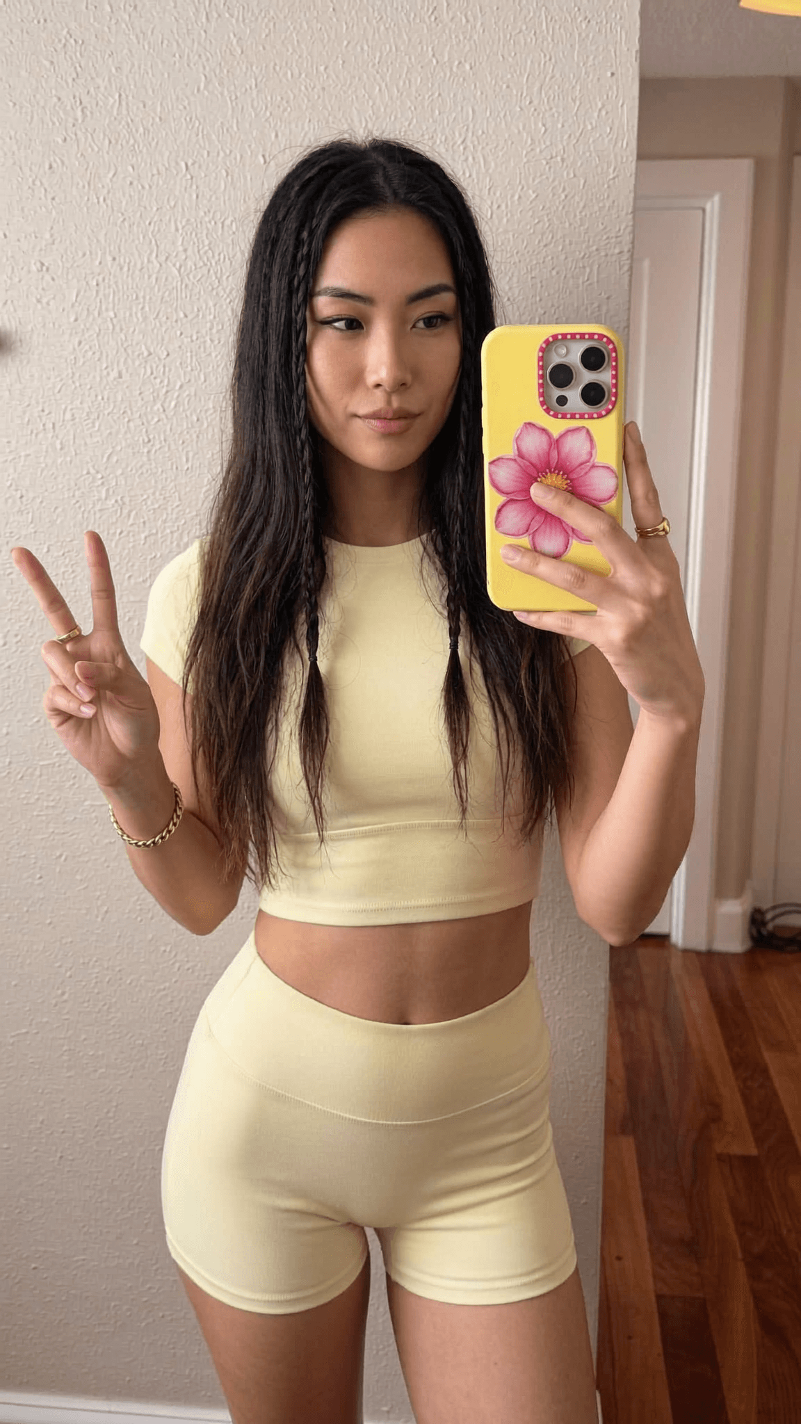 A woman in a yellow crop top and shorts poses for a mirror selfie, making a peace sign with her hand.