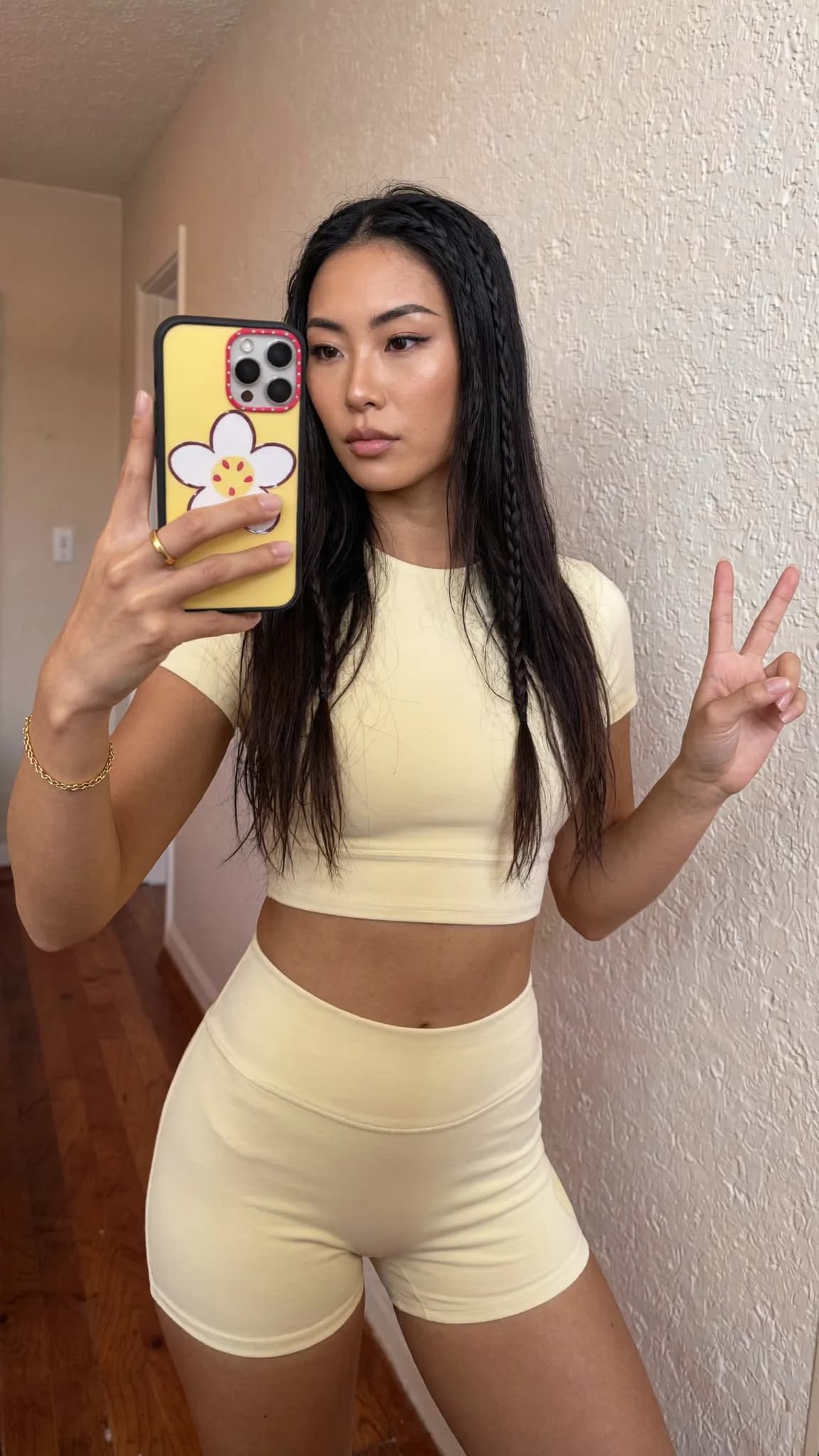 A woman in a yellow crop top and shorts poses for a mirror selfie, making a peace sign with her hand.