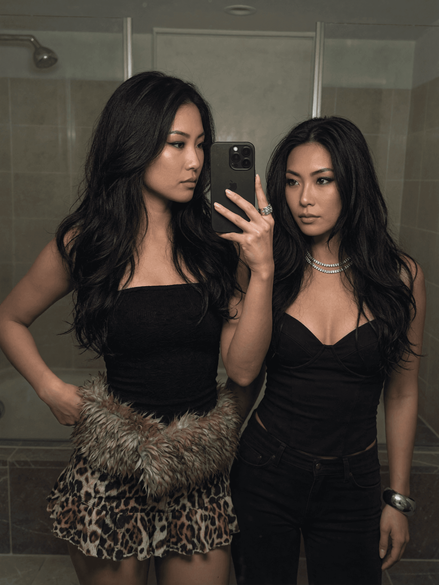 Two individuals in a bathroom taking a mirror selfie, dressed casually with one wearing a leopard print skirt and the other in black attire.