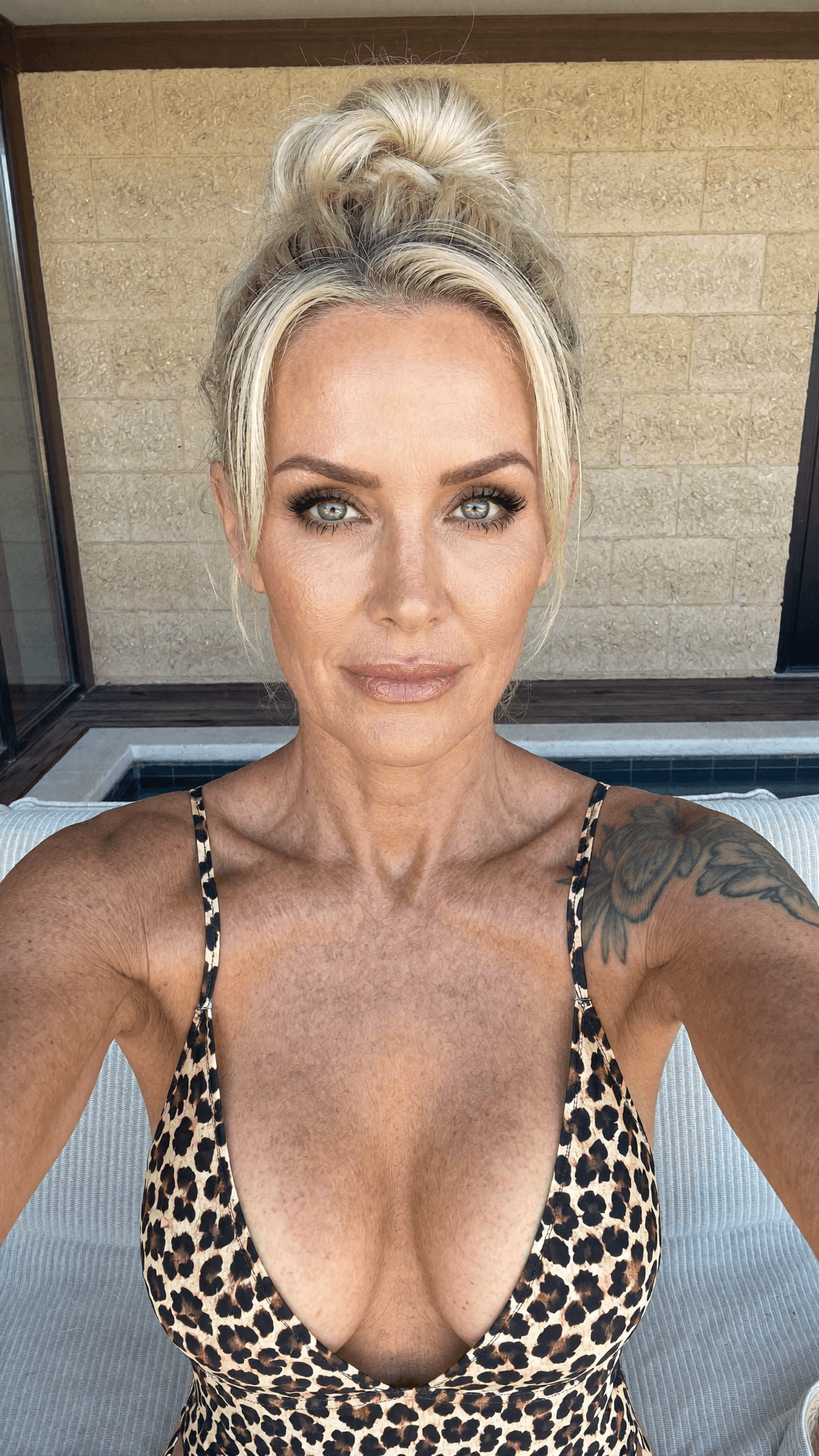 A woman with a blonde updo and leopard print swimsuit poses for the camera outdoors.