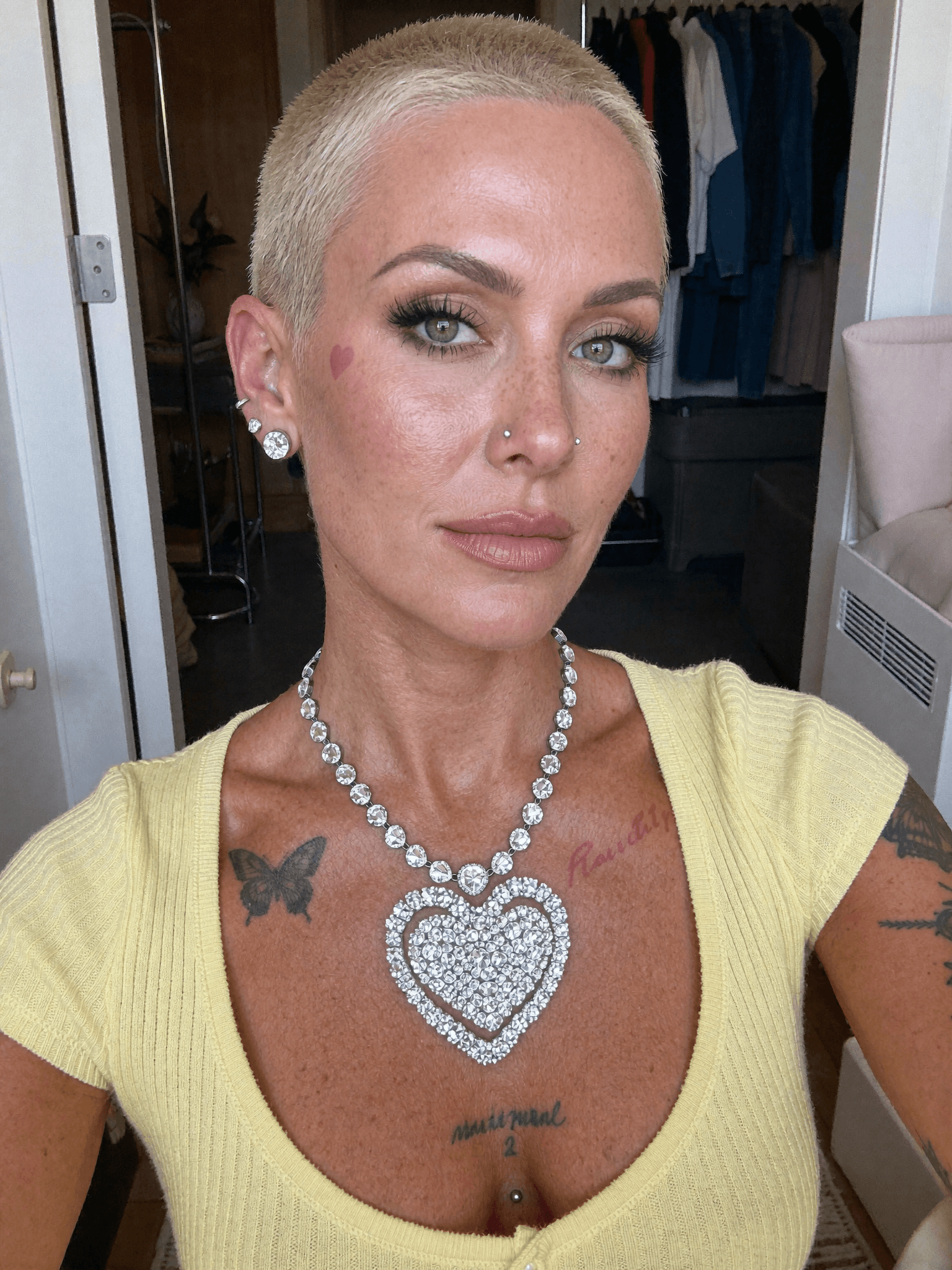 A woman with a platinum blonde pixie cut and a septum ring poses in a bedroom, wearing casual clothing.