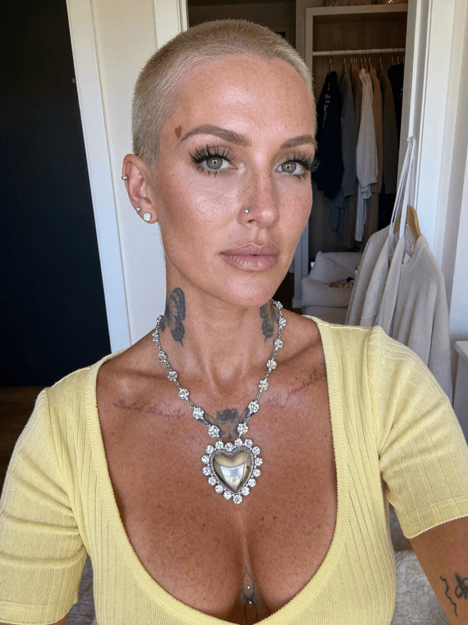 A woman with a platinum blonde pixie cut and septum ring poses in a bedroom, wearing casual clothing.