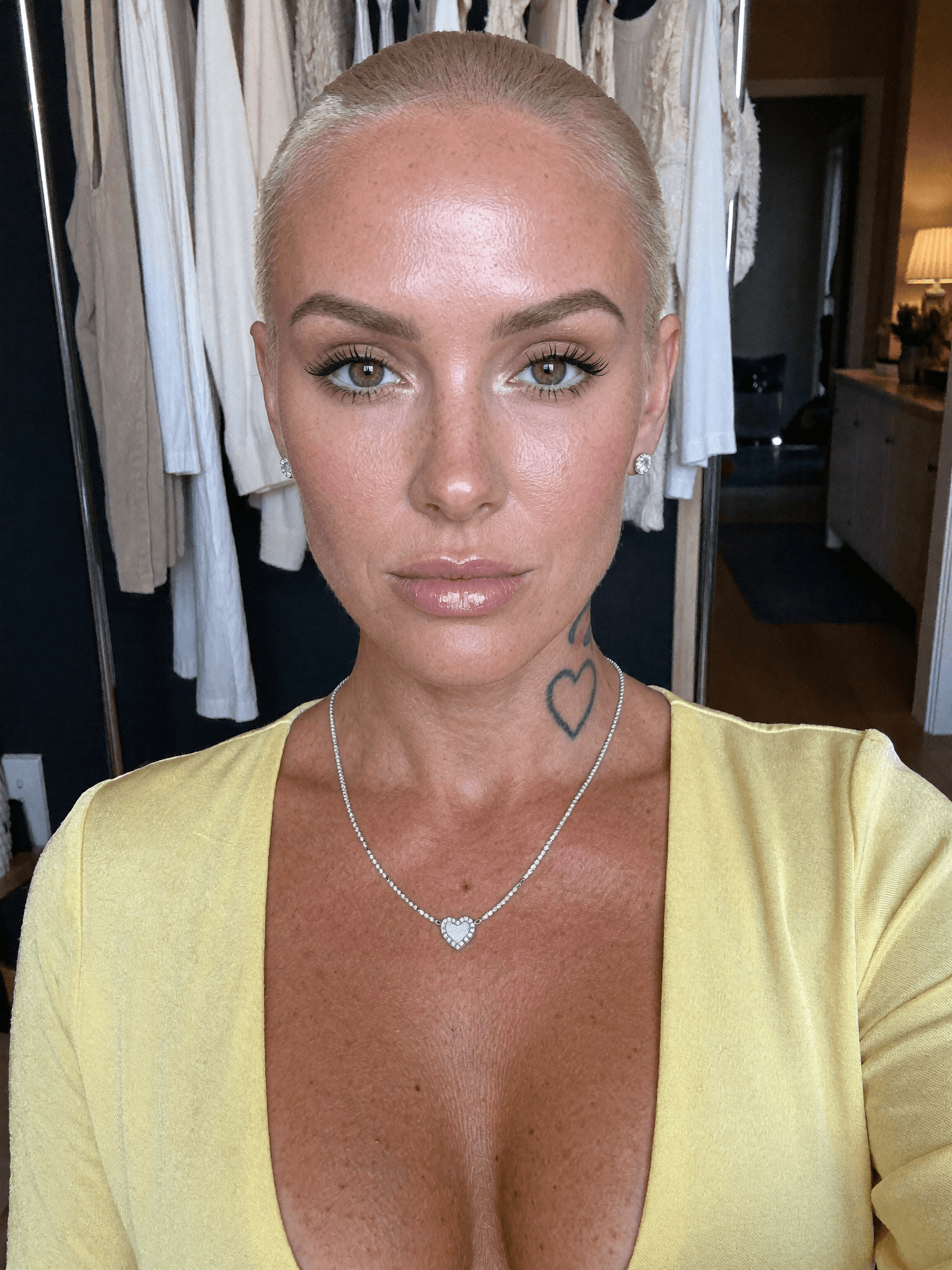 A woman with a platinum blonde pixie cut poses in a bedroom, wearing casual clothing and jewelry.