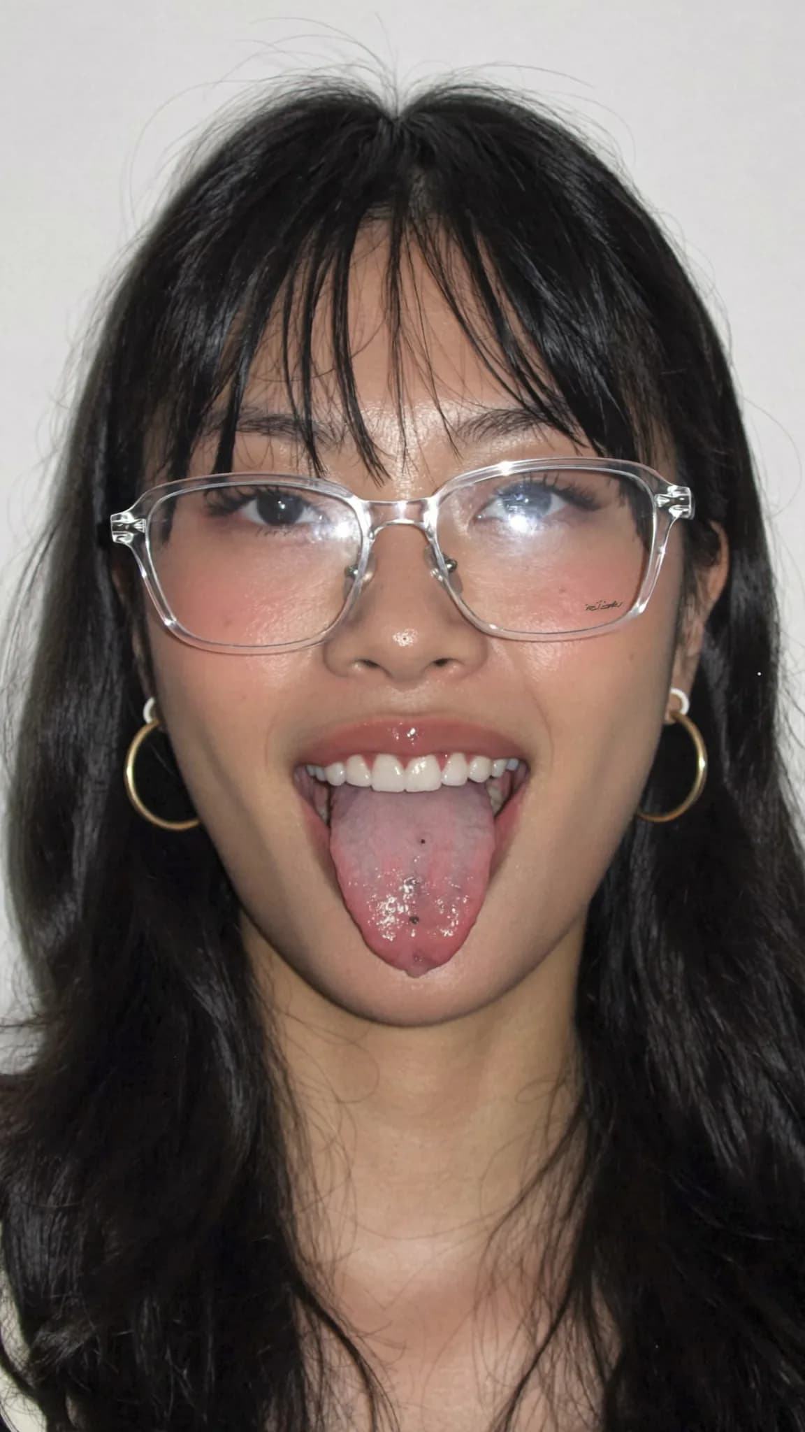 A young woman with long black hair and glasses is sticking out her tongue in a playful pose.
