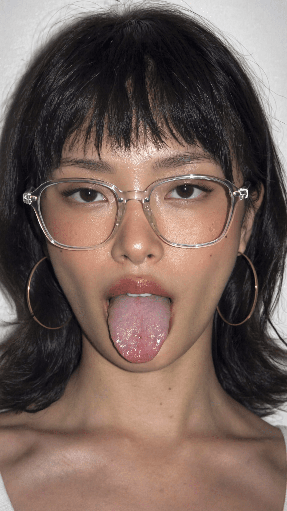 A young woman with short black hair and glasses is sticking out her tongue in a playful pose.