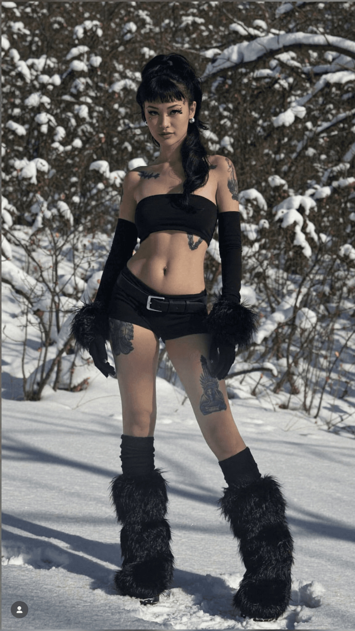 A woman stands confidently in a snowy outdoor setting, wearing semi-nude attire with bold tattoos and fur accessories.