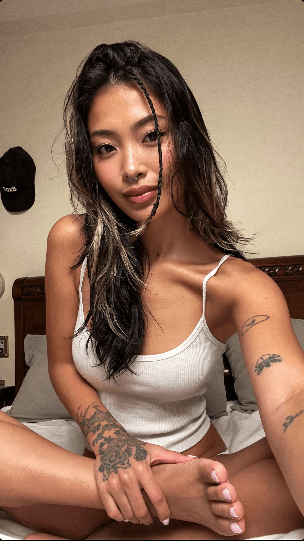 A woman with a septum ring and tattoos sits in an intimate pose on a bed.
