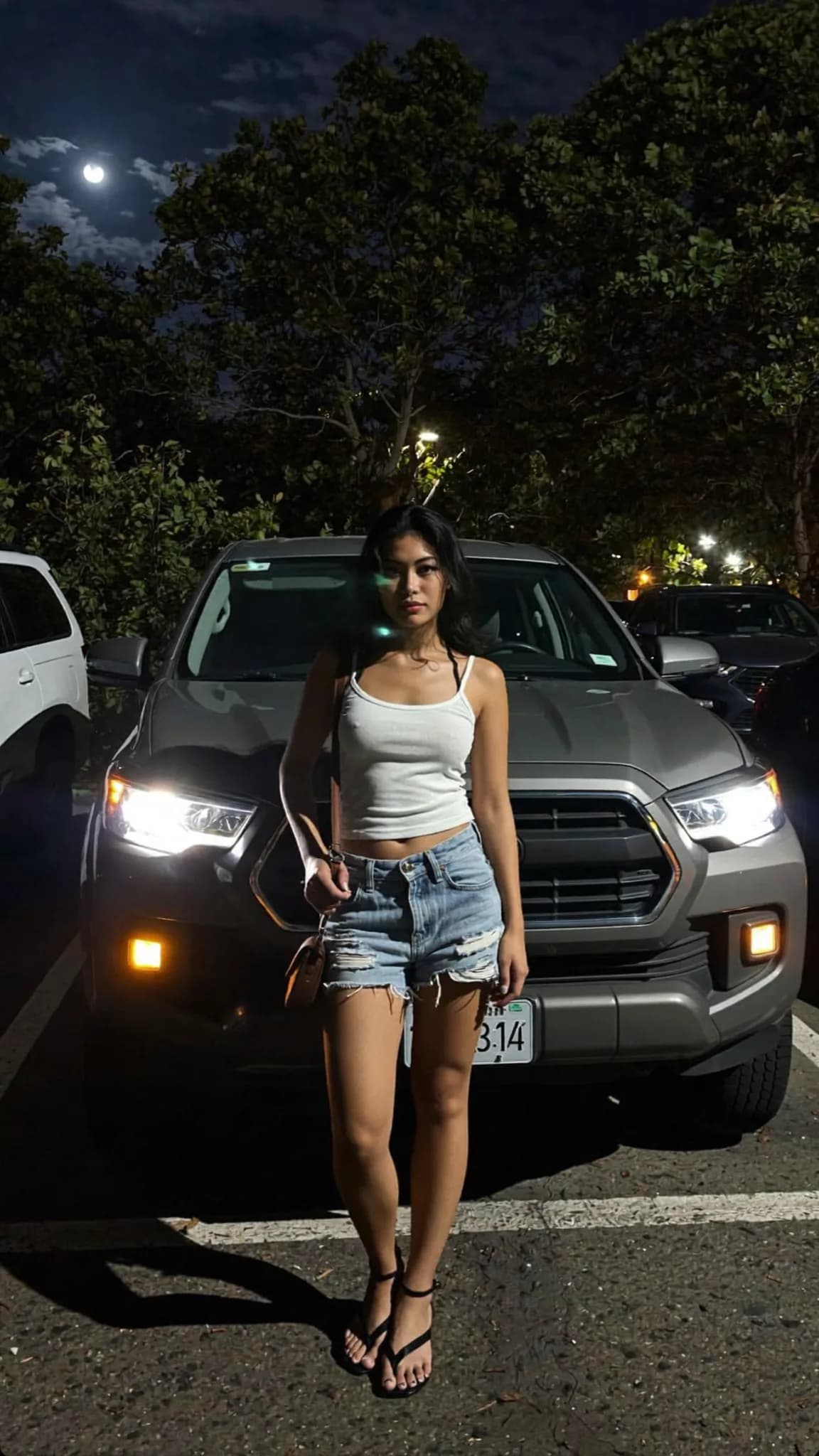 A woman stands confidently in front of a car at night, wearing casual clothing and exuding a playful mood.