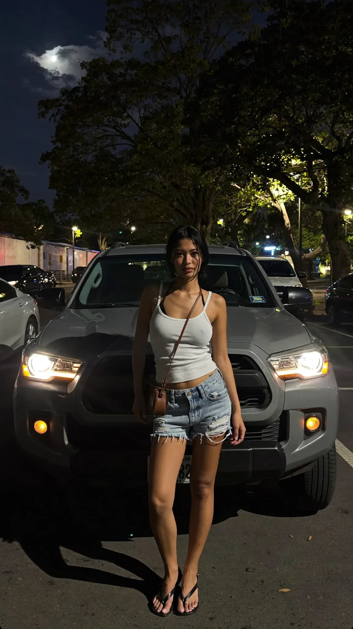 A woman stands confidently in front of a car at night, wearing casual clothing and exuding a playful mood.