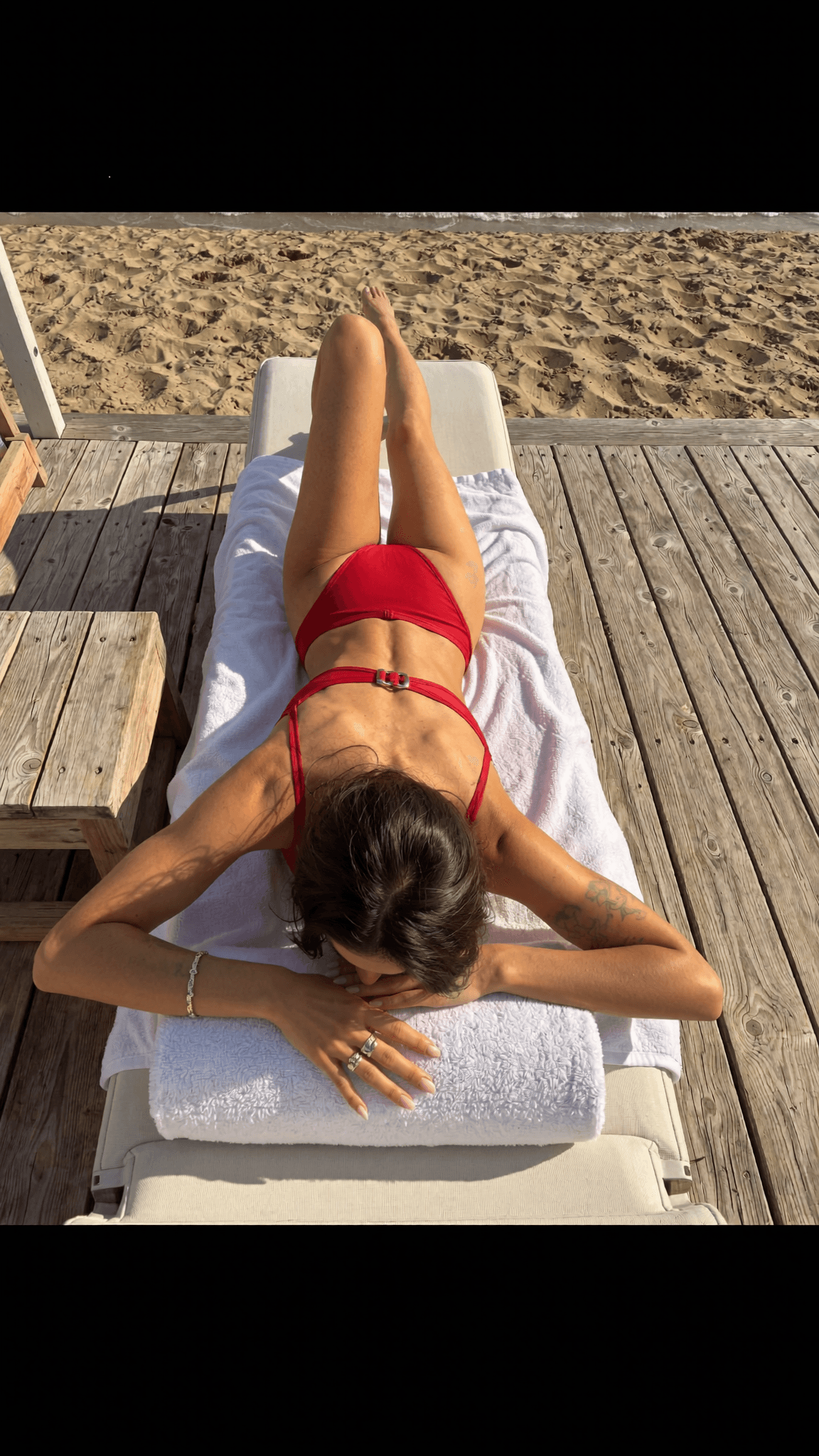 A woman in a red swimsuit lies on her stomach on a beach chair, facing the sand.