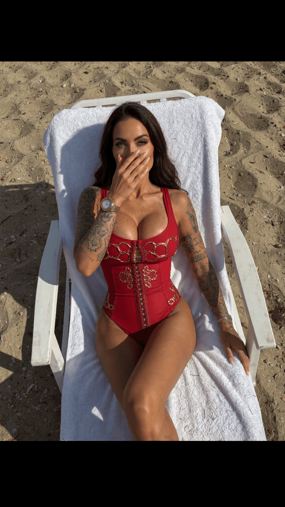 A woman in a red swimsuit sits on a beach chair, covering her mouth with one hand.