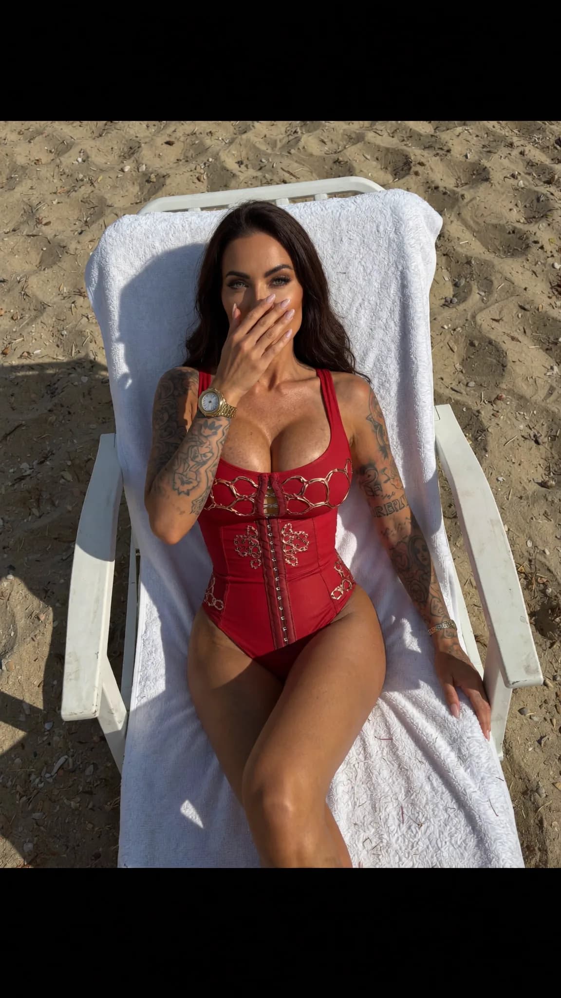 A woman in a red swimsuit sits on a beach chair, covering her mouth with one hand.