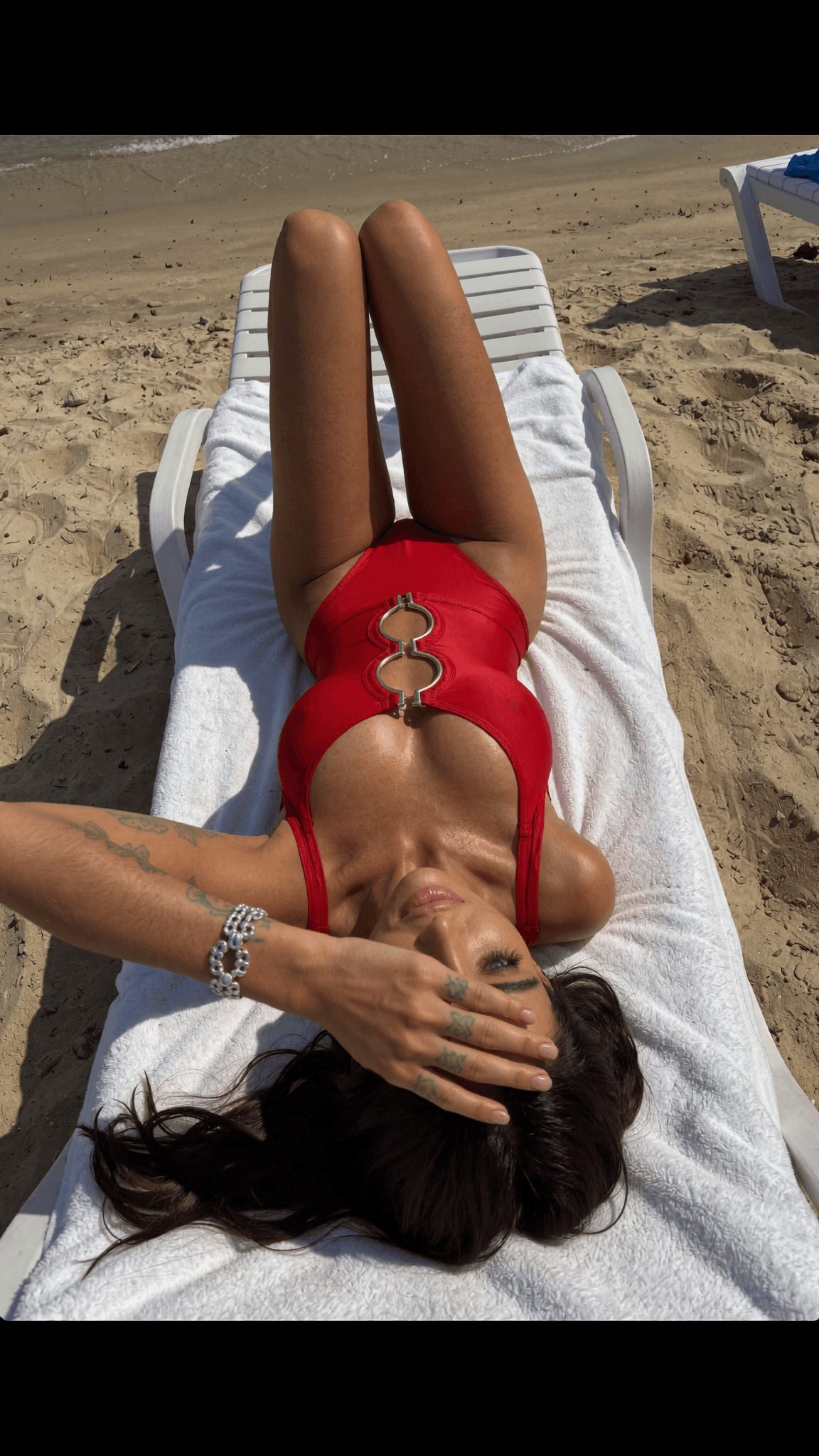 A woman in a red swimsuit lies on her stomach on the beach, wearing jewelry and with tattoos visible.