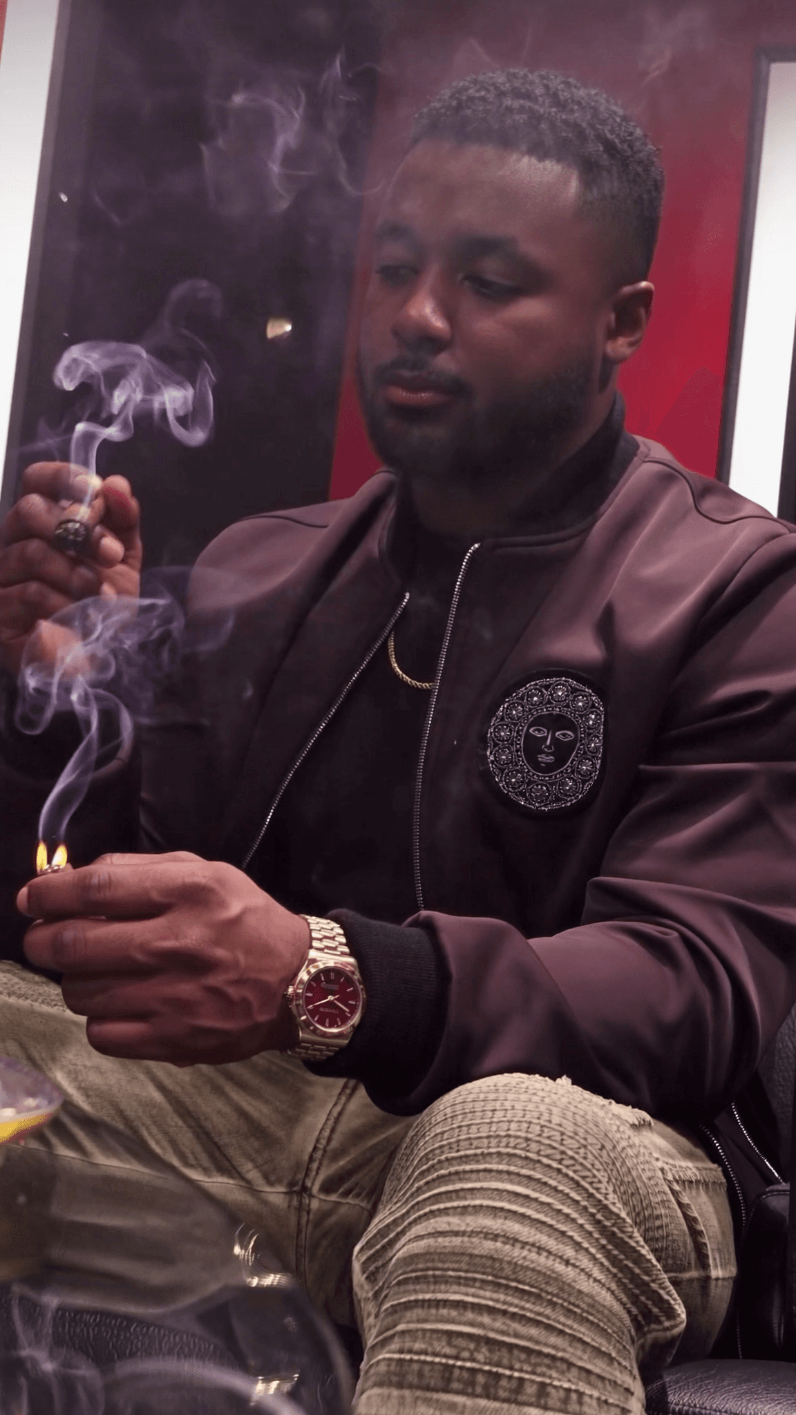 A man in casual attire is seated, lighting a cigar with smoke rising around him.