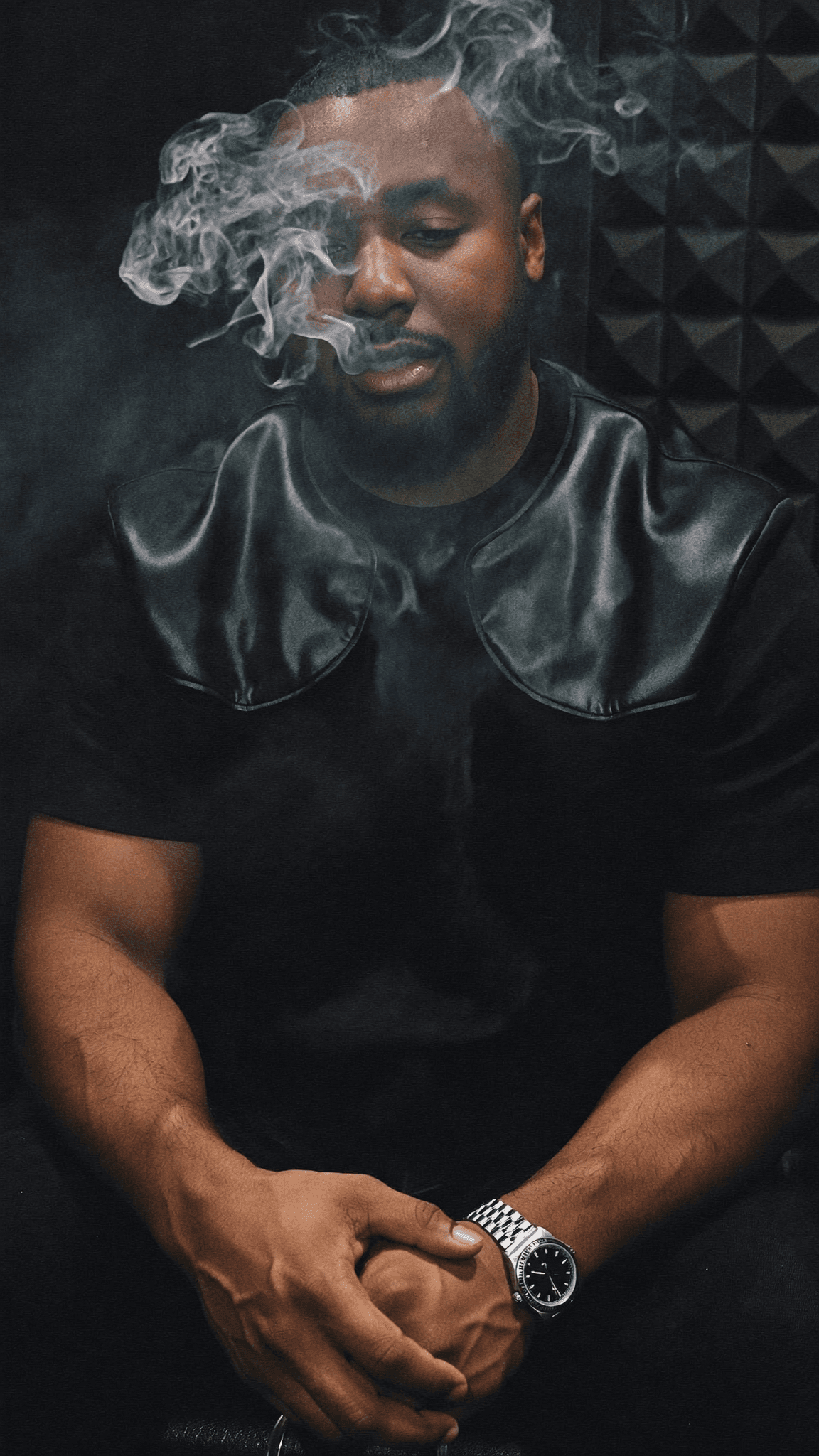 A man in casual attire sits with crossed hands, partially obscured by smoke.