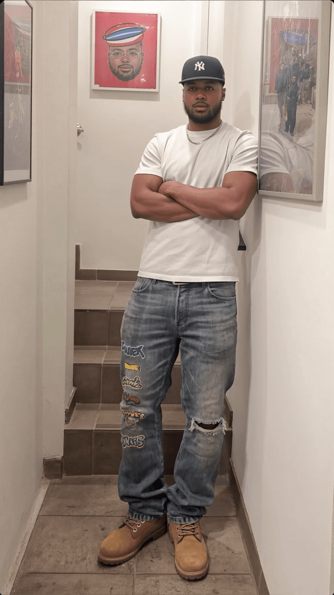 A man stands in a hallway wearing casual clothing with his arms crossed.