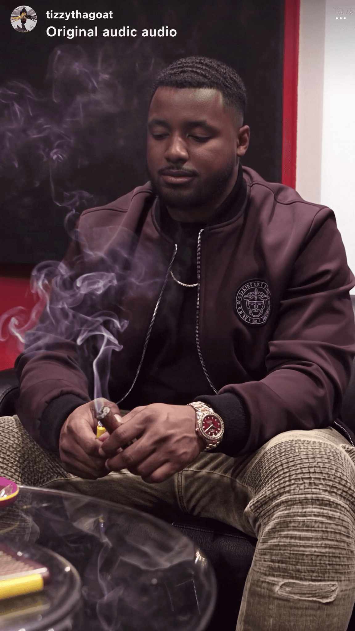 A man in casual attire is seated, smoking a cigar with smoke rising around him.
