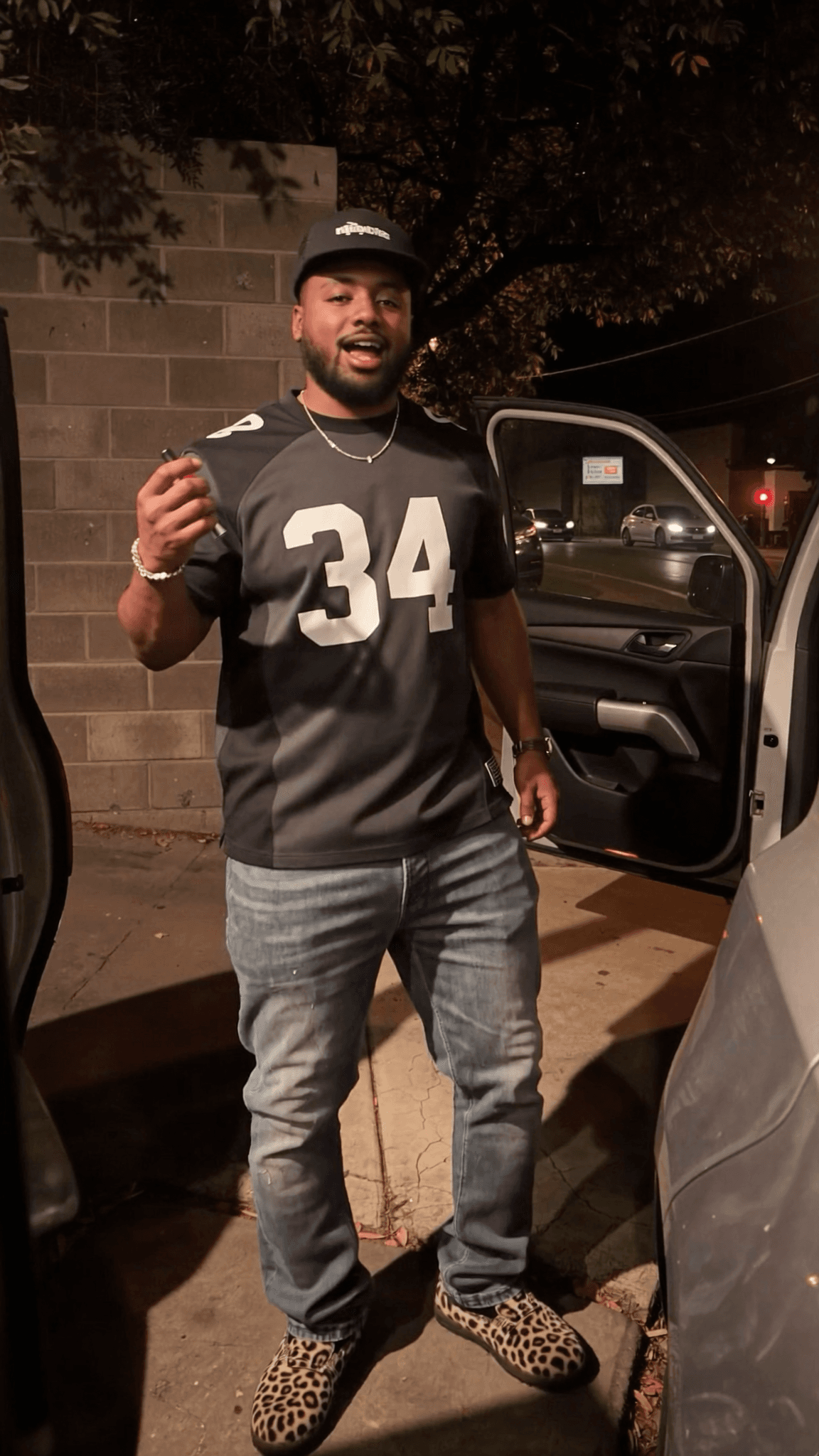 A man stands next to an open car door wearing a sports jersey and casual jeans.