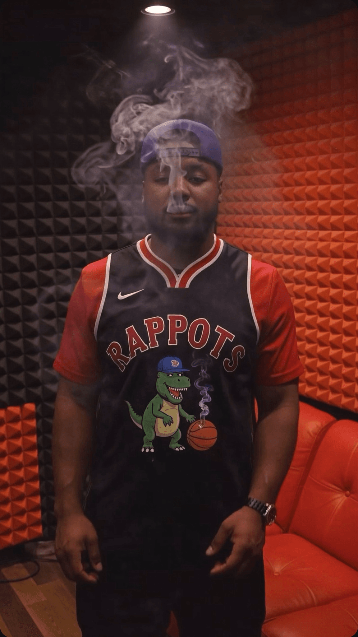 A person wearing a basketball jersey with a cartoon dinosaur graphic is standing in a studio, exhaling smoke.