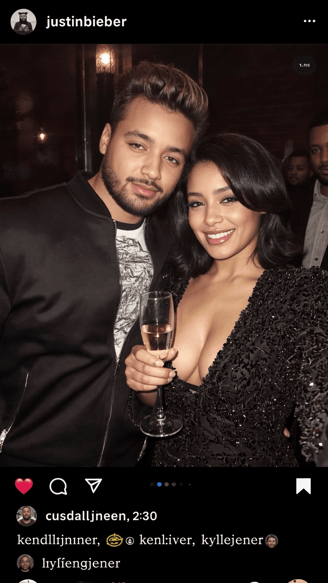 A woman in a formal dress and a man in casual attire pose together at a club, holding champagne glasses.