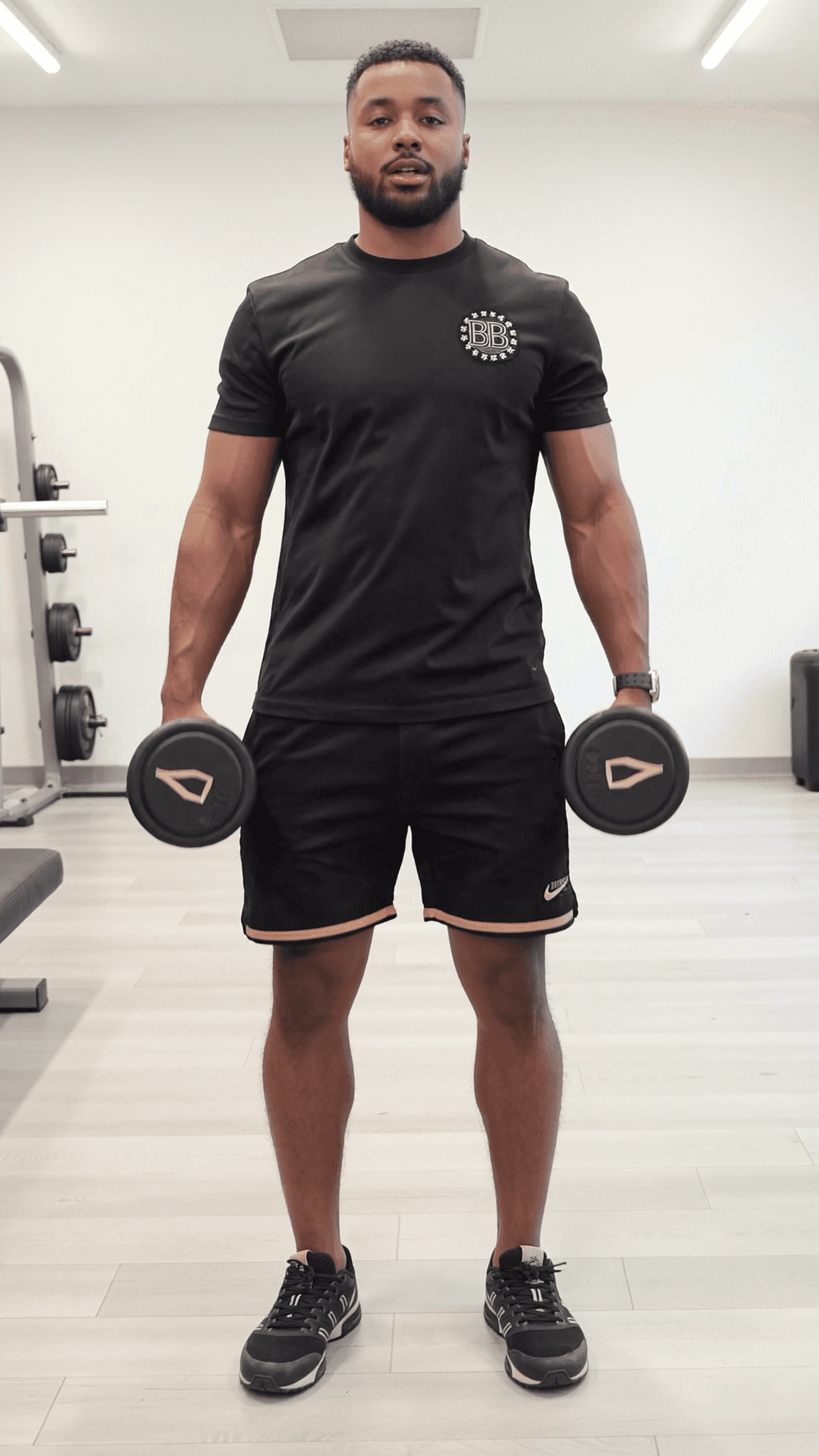 A muscular man in a black athletic outfit holds dumbbells while standing confidently in a studio.