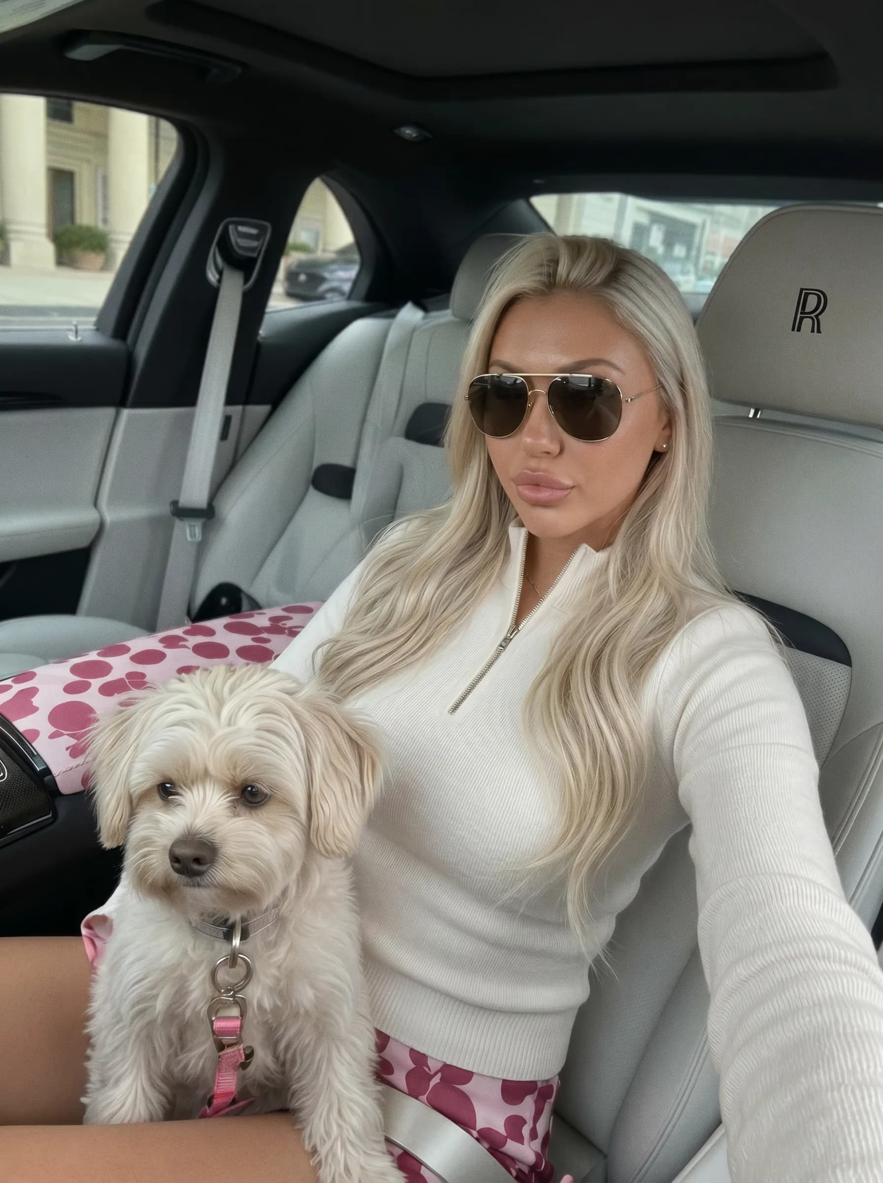 A woman with long blonde hair and sunglasses sits in a car, holding a small white dog.