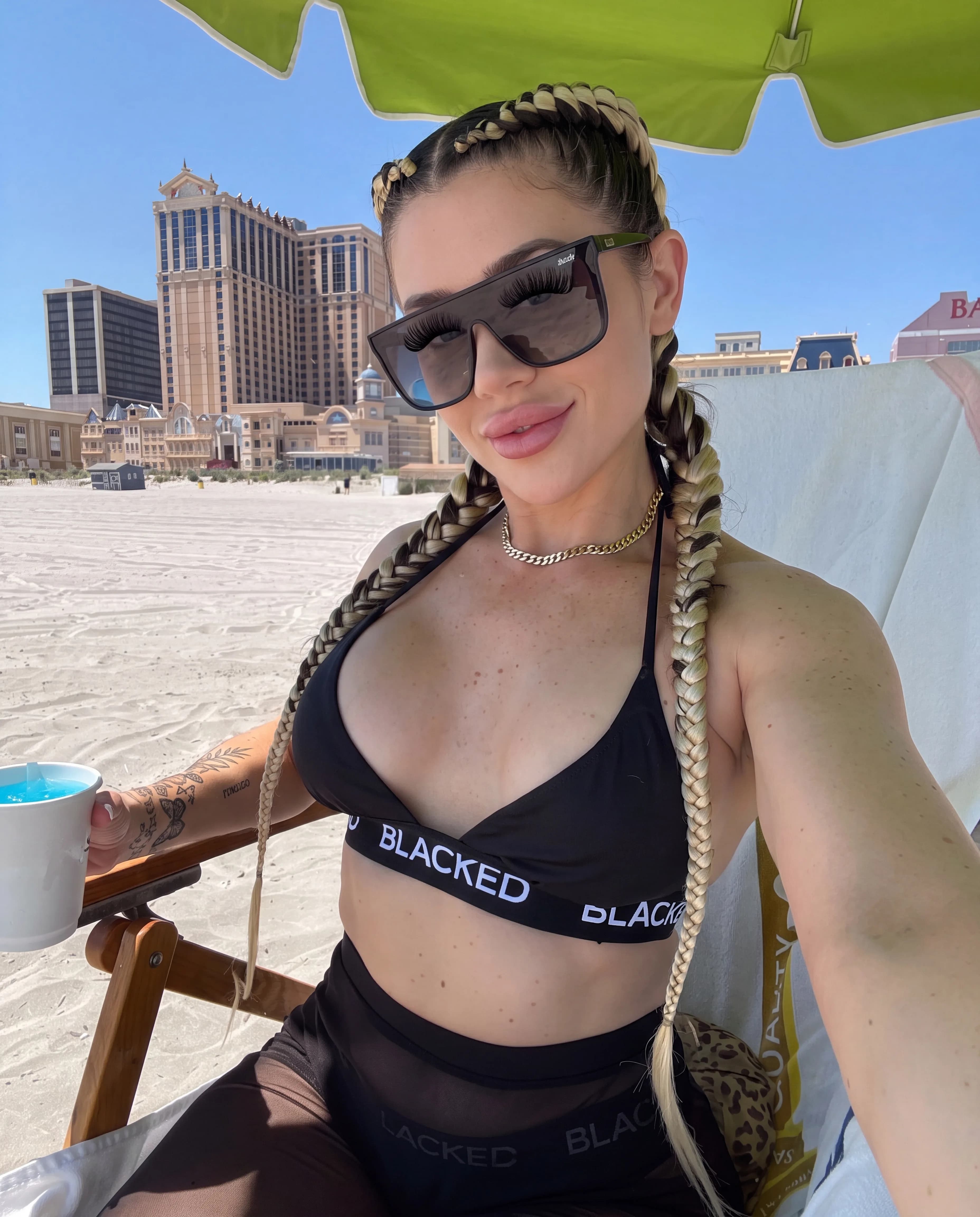 A woman with blonde braids and a black swimsuit sits on the beach under an umbrella, holding a drink.