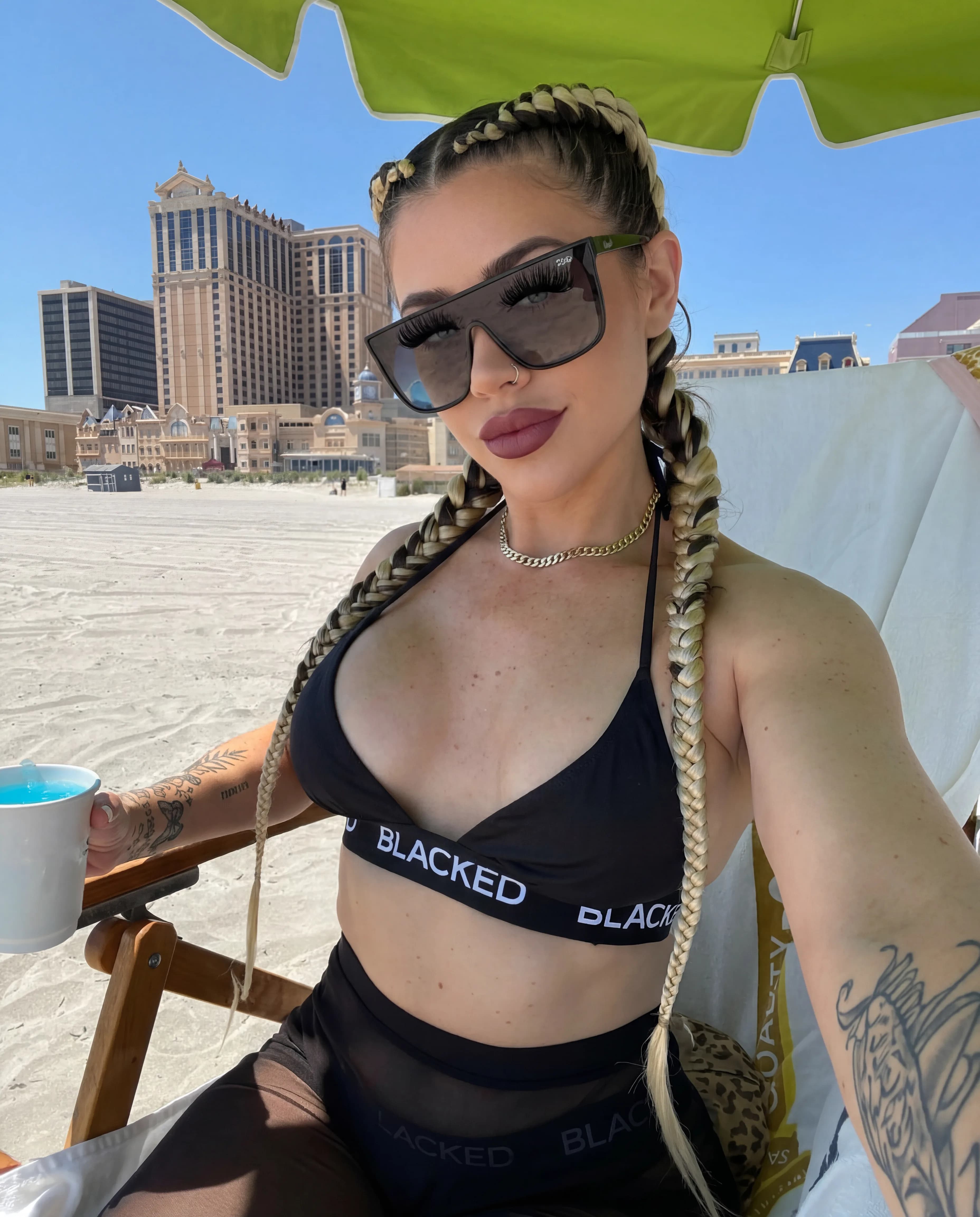 A woman with blonde braids and a black swimsuit sits on the beach under an umbrella, holding a drink.