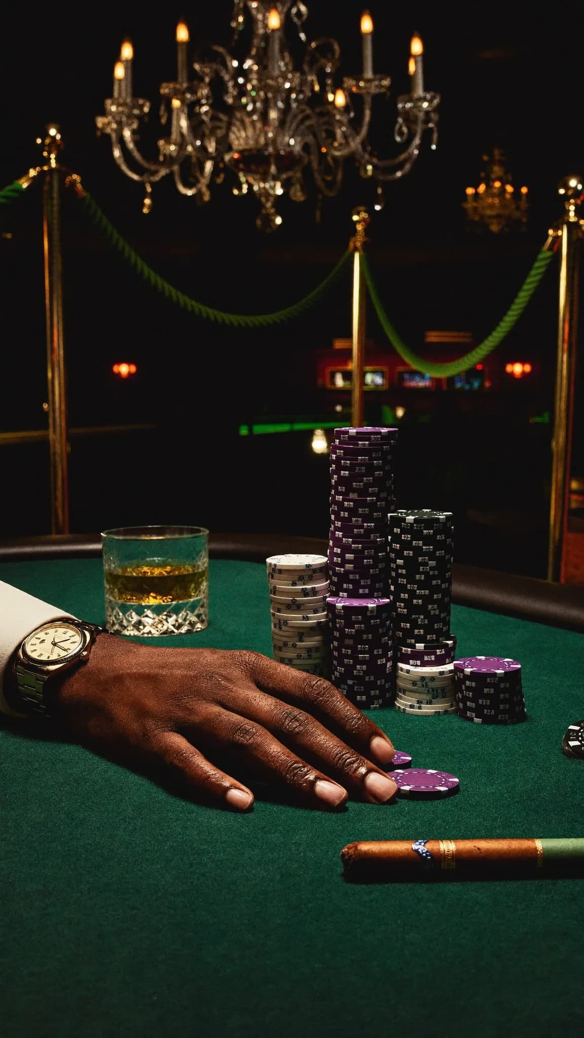 A person is seated at a poker table with stacks of chips and a glass of whiskey, wearing a watch.
