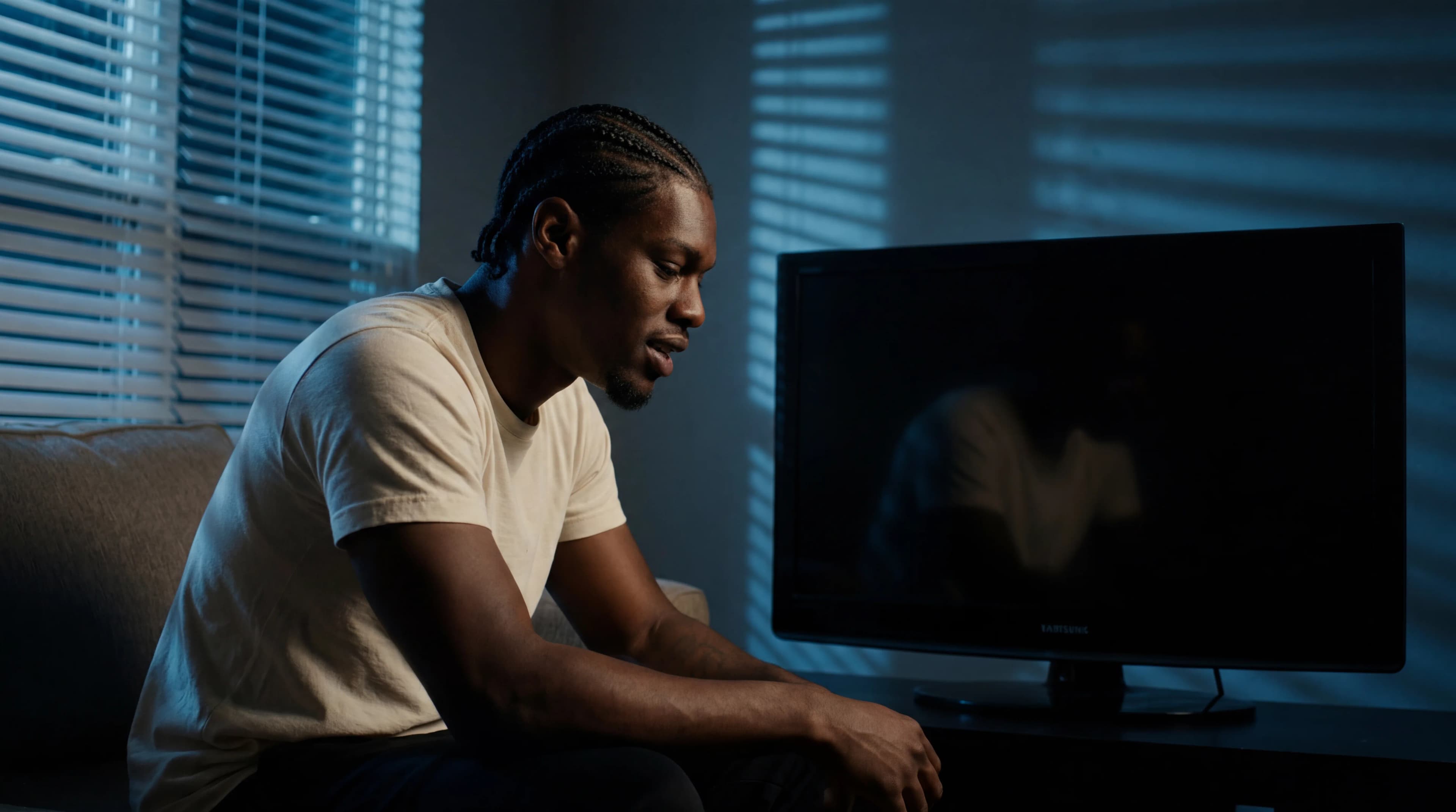 A man in a casual outfit is seated and looking at a TV screen, with soft lighting casting shadows on the wall.