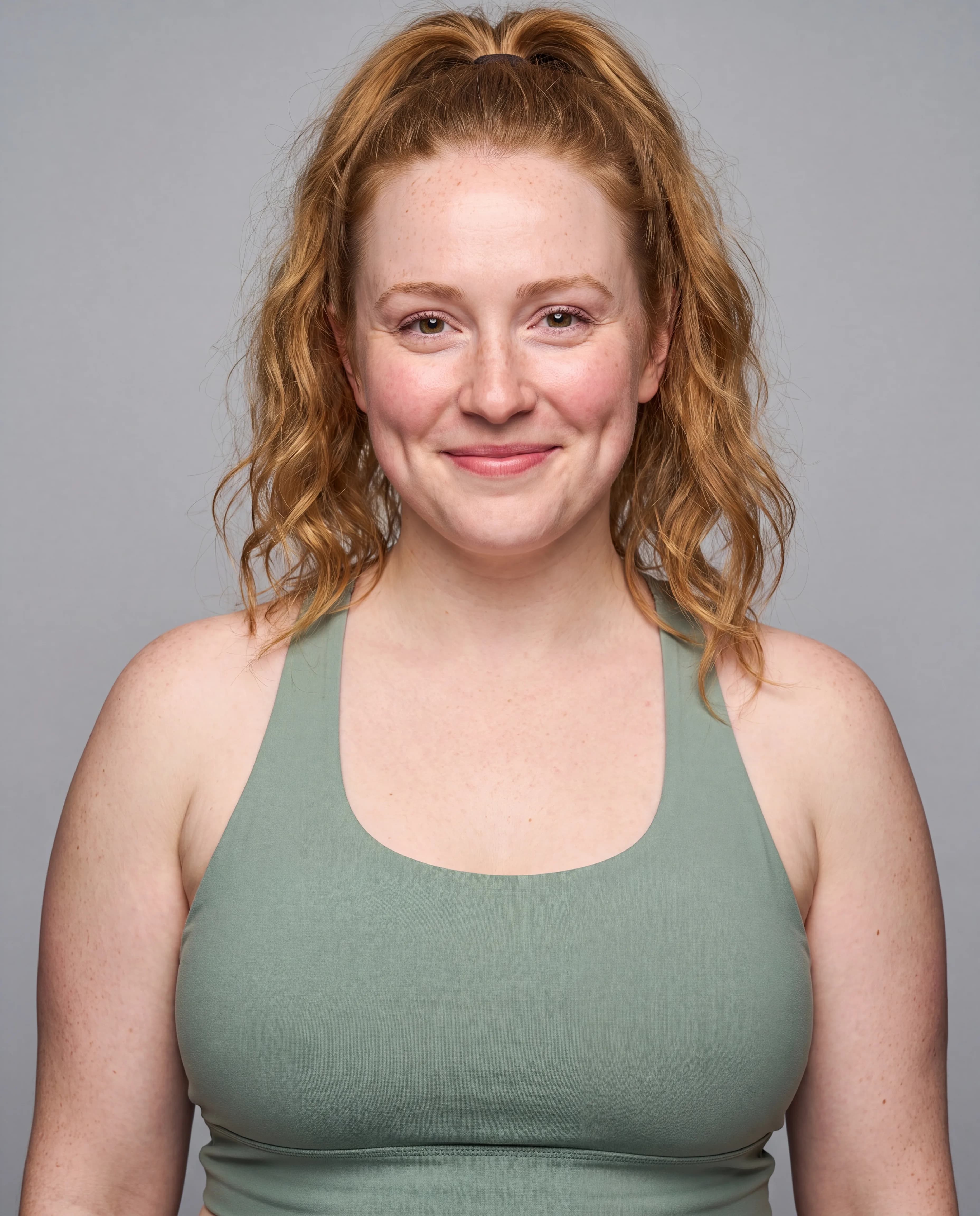 A woman with curly red hair wearing a green tank top is posing in a studio setting.