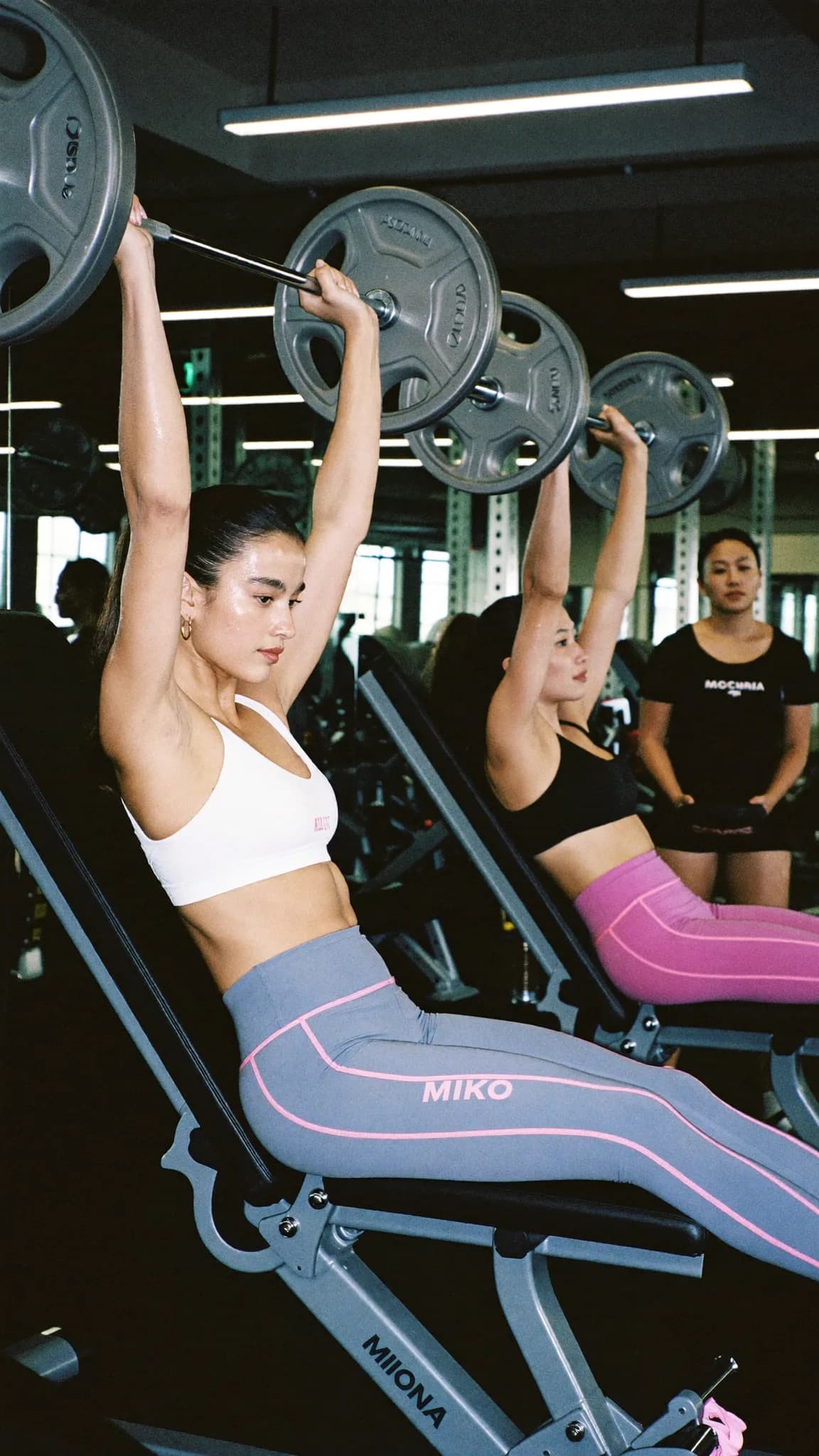 Two women in athletic wear perform seated weightlifting exercises in a gym setting.