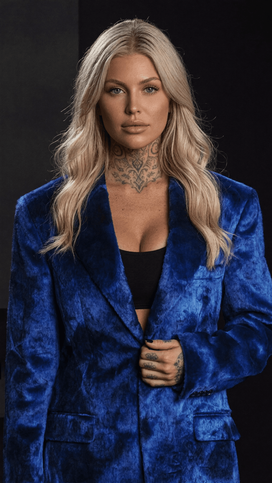 A woman with platinum blonde hair and a blue velvet blazer poses confidently in a studio setting.
