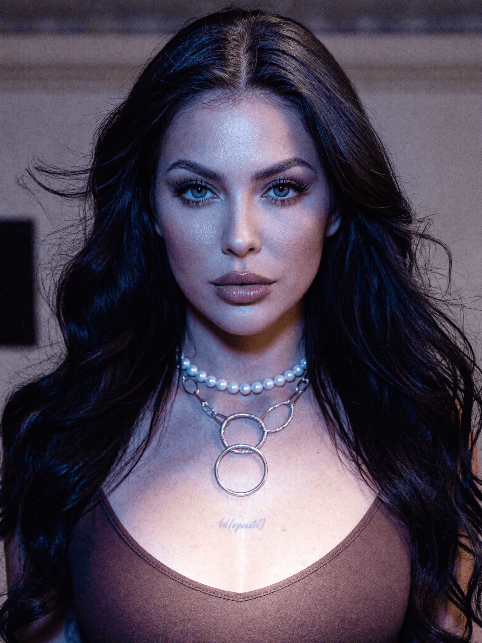 A woman with long black hair and striking blue eyes poses in a studio setting, wearing casual clothing and jewelry.